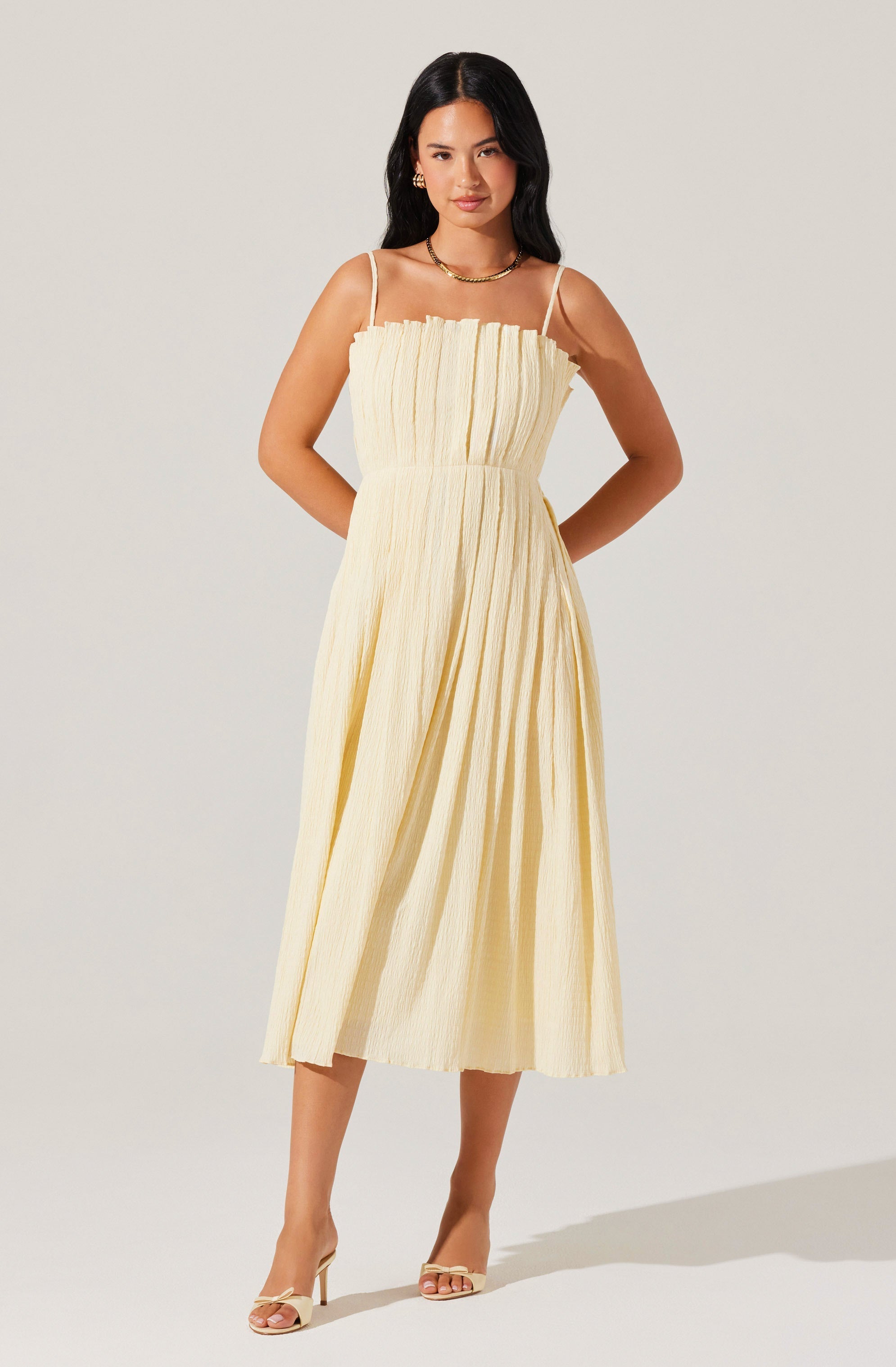 A woman is wearing a light yellow, sleeveless, ankle-length dress with thin spaghetti straps. The dress features a gathered, pleated bodice and a full, flowing skirt with subtle vertical stripes. She has accessorized with gold jewelry, including hoop earrings and a chain necklace, and is wearing open-toed, beige high-heel sandals.