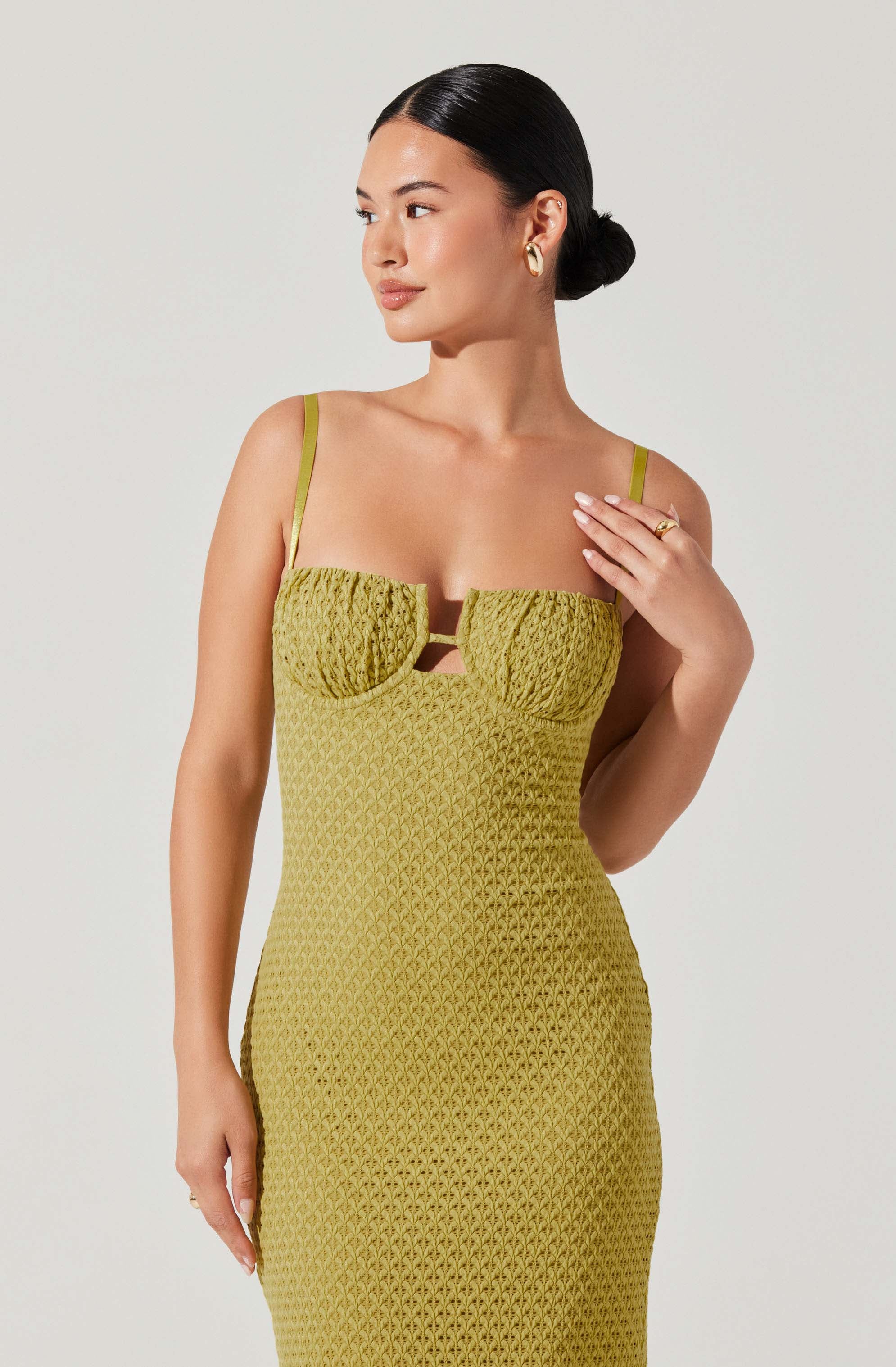 A woman is wearing a fitted, sleeveless, olive-green dress with thin straps. The dress features textured, ruched fabric on the bust area with a small cut-out detail beneath the chest. The lower part of the dress has a diamond-shaped pattern. She has her dark hair styled in a sleek bun and is accessorized with gold earrings and a gold ring on her finger.