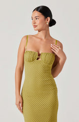 A woman is wearing a fitted, sleeveless, olive-green dress with thin straps. The dress features textured, ruched fabric on the bust area with a small cut-out detail beneath the chest. The lower part of the dress has a diamond-shaped pattern. She has her dark hair styled in a sleek bun and is accessorized with gold earrings and a gold ring on her finger.