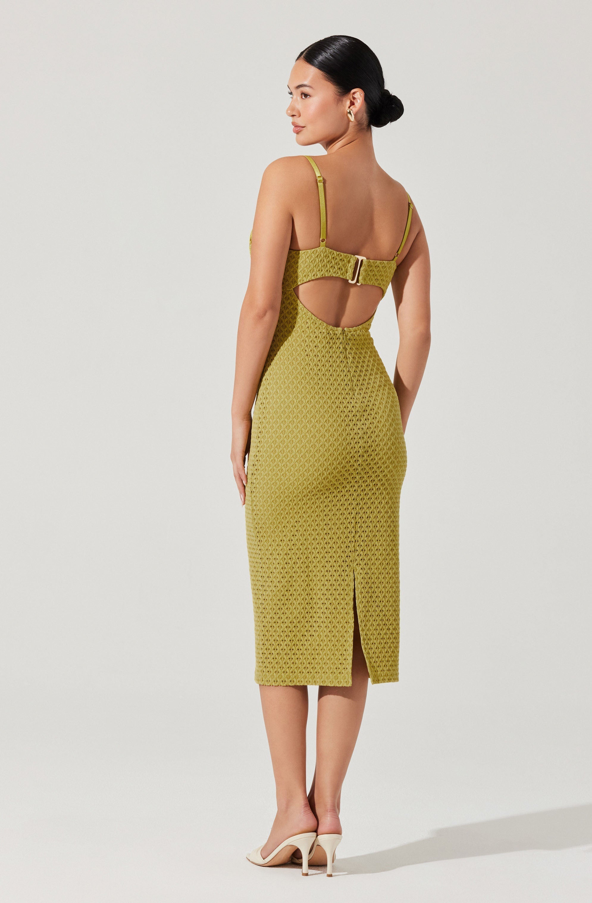 A woman wearing a fitted, sleeveless dress in a green geometric pattern. The dress features adjustable shoulder straps, a wide waistband with a metallic buckle, and a cutout detail at the back with a small slit at the hem. She is also wearing light-colored high-heeled shoes.
