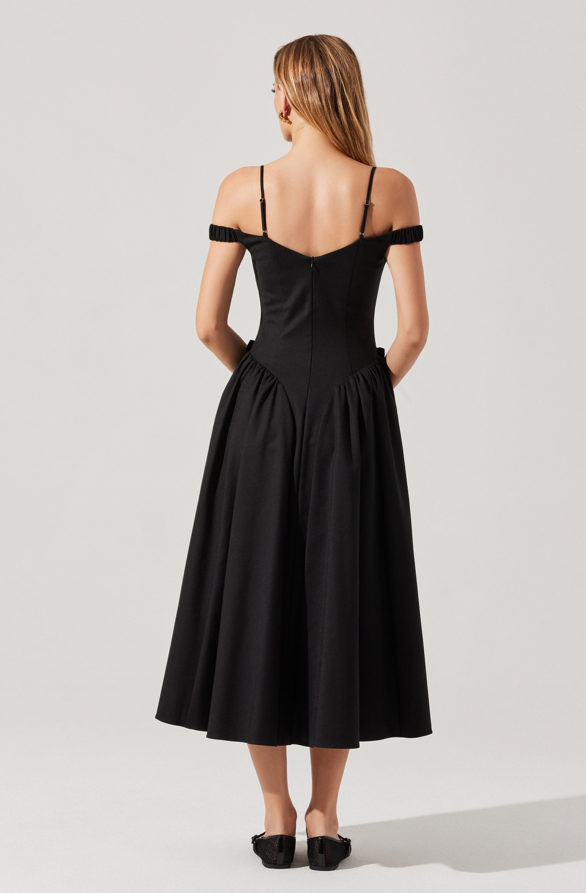 A woman is wearing a black off-the-shoulder dress with thin spaghetti straps and ruffle details on the sleeves. The dress has a fitted bodice and a full, gathered skirt that falls to mid-calf length. She is also wearing black shoes with a textured pattern.