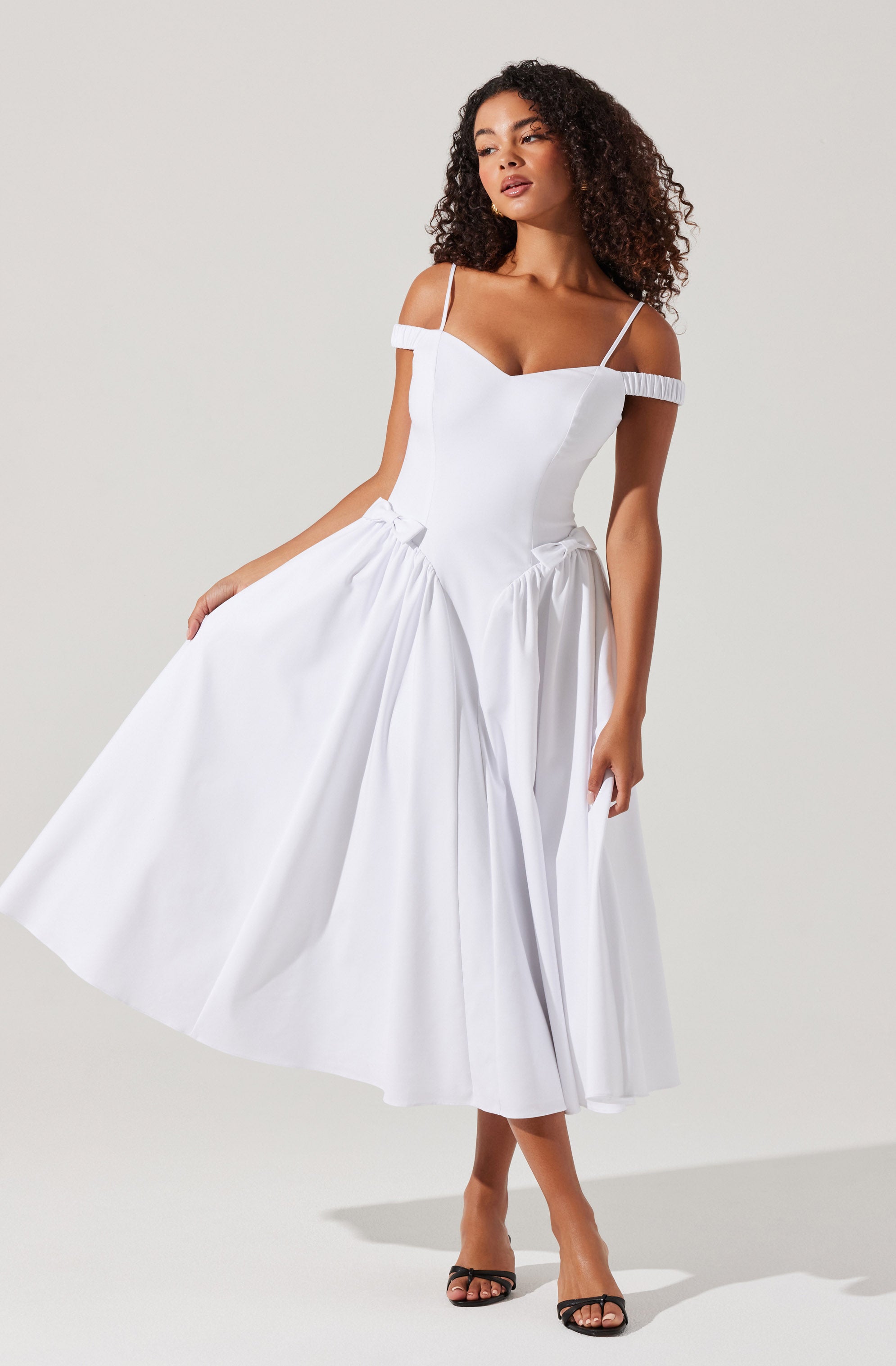 A woman is wearing a white dress with off-the-shoulder sleeves and thin spaghetti straps. The dress has a fitted bodice and a full, pleated skirt that extends to mid-calf length. The fabric appears lightweight and has a subtle sheen.