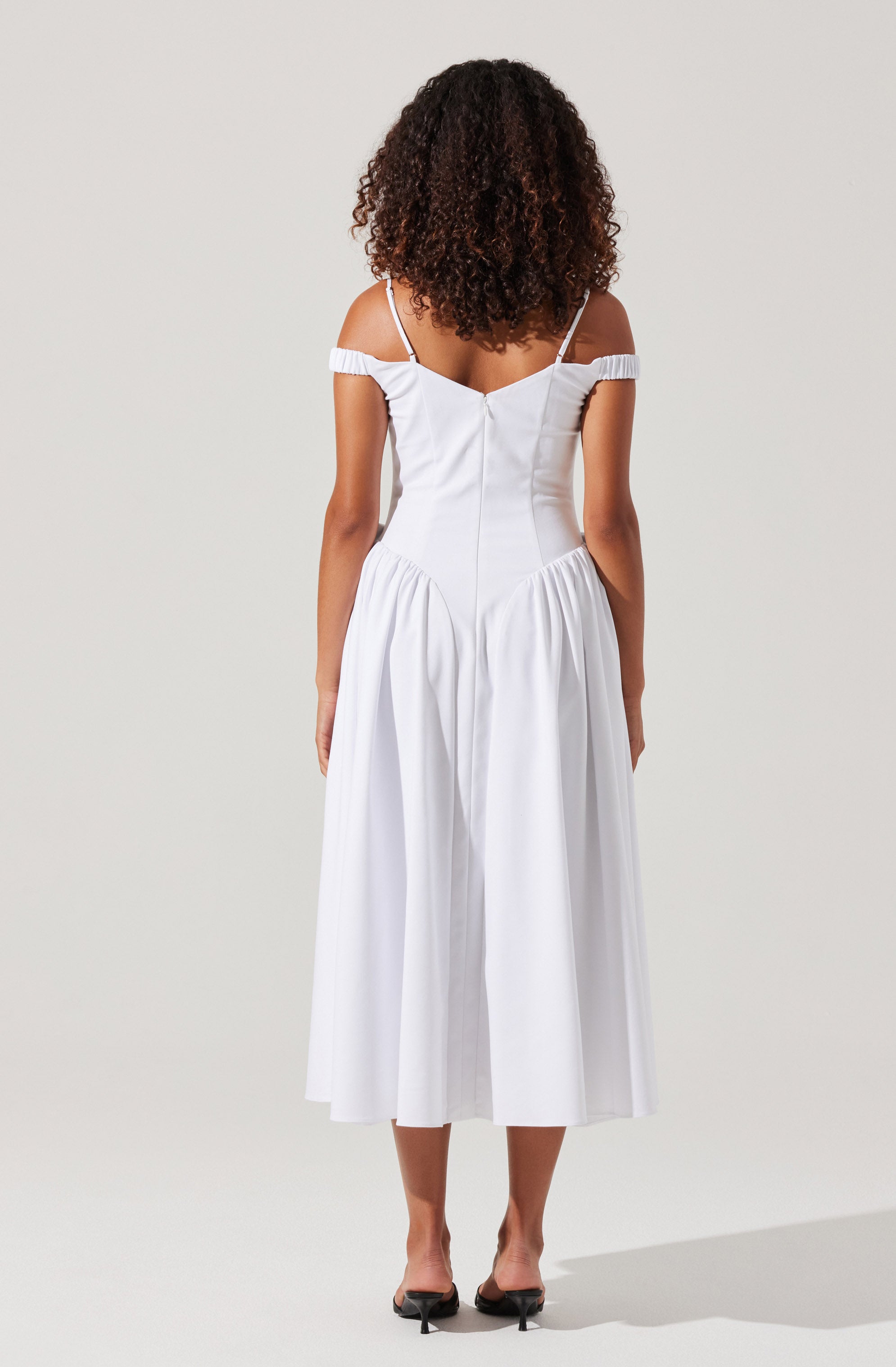 A woman wearing a white, off-the-shoulder dress with thin spaghetti straps. The dress features a fitted bodice and a full, gathered skirt that falls below the knees. She is also wearing black, open-toe heels.