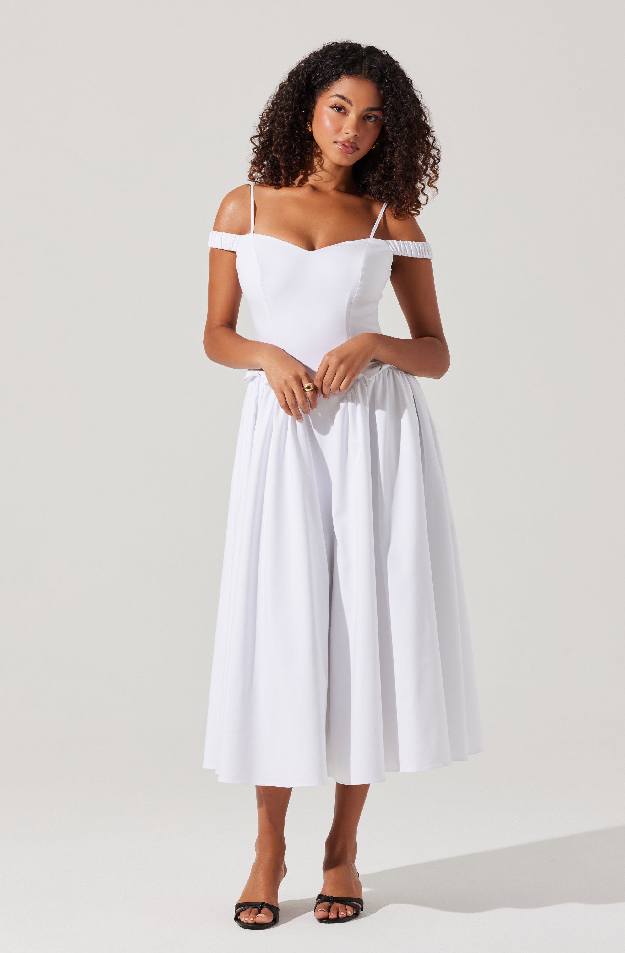 A woman is wearing a white dress with thin spaghetti straps and off-the-shoulder sleeves that have a textured band. The dress has a fitted bodice and a high-waisted, wide, pleated skirt that falls to mid-calf length. She is also wearing black open-toe sandals with ankle straps.