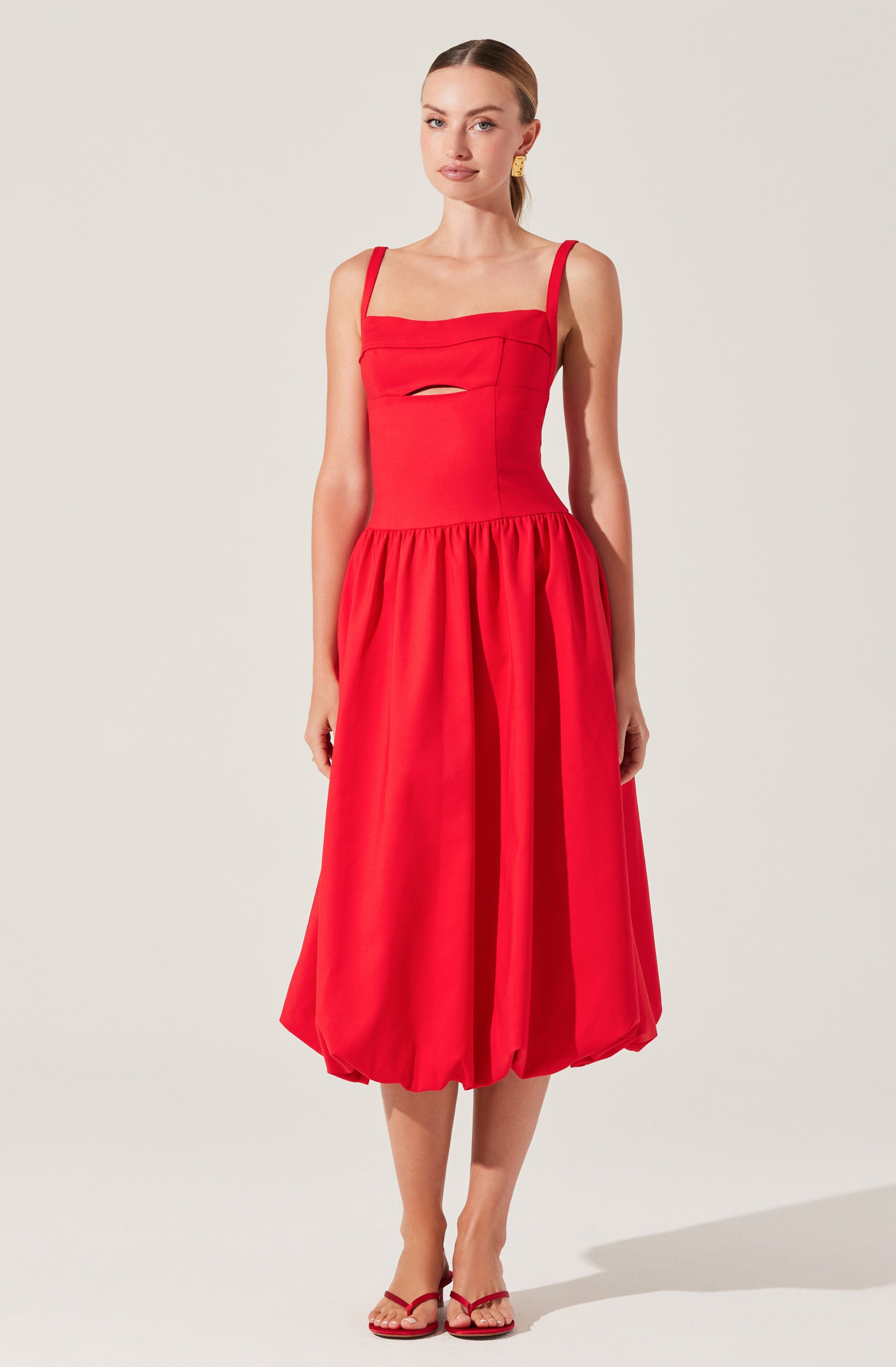A woman is wearing a sleeveless red dress with thin shoulder straps. The bodice features a horizontal seam across the chest with a small cutout detail just below it. The skirt is voluminous with gathered fabric that creates a bubble hemline ending below the knees. She has paired the dress with red and beige open-toe high heels and is wearing gold earrings.