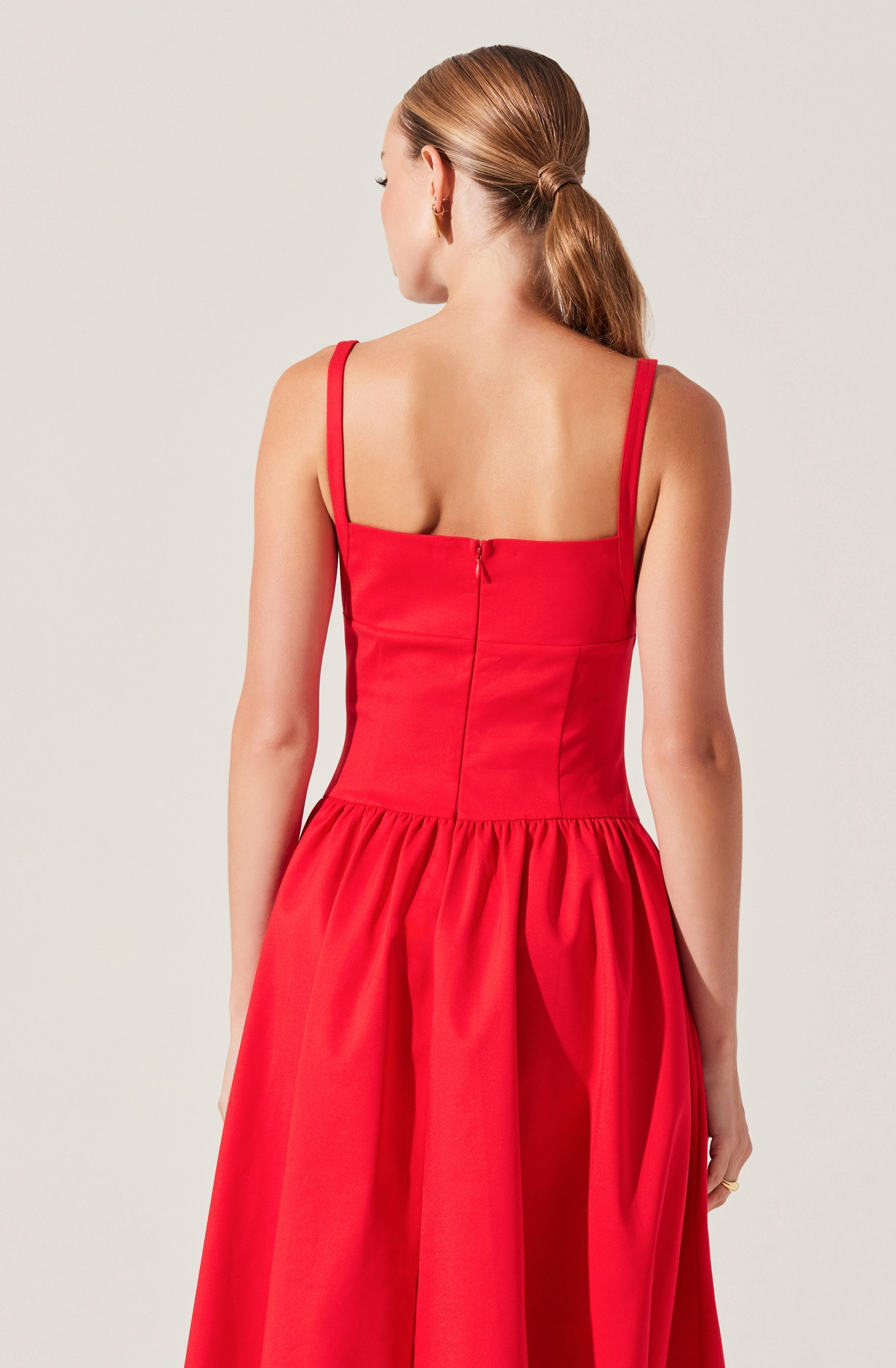 A woman is wearing a sleeveless, red dress with fitted bodice and thin shoulder straps. The back of the dress features a zipper closure and a gathered, full skirt that extends below the waist. The fabric of the dress appears smooth and slightly shiny.