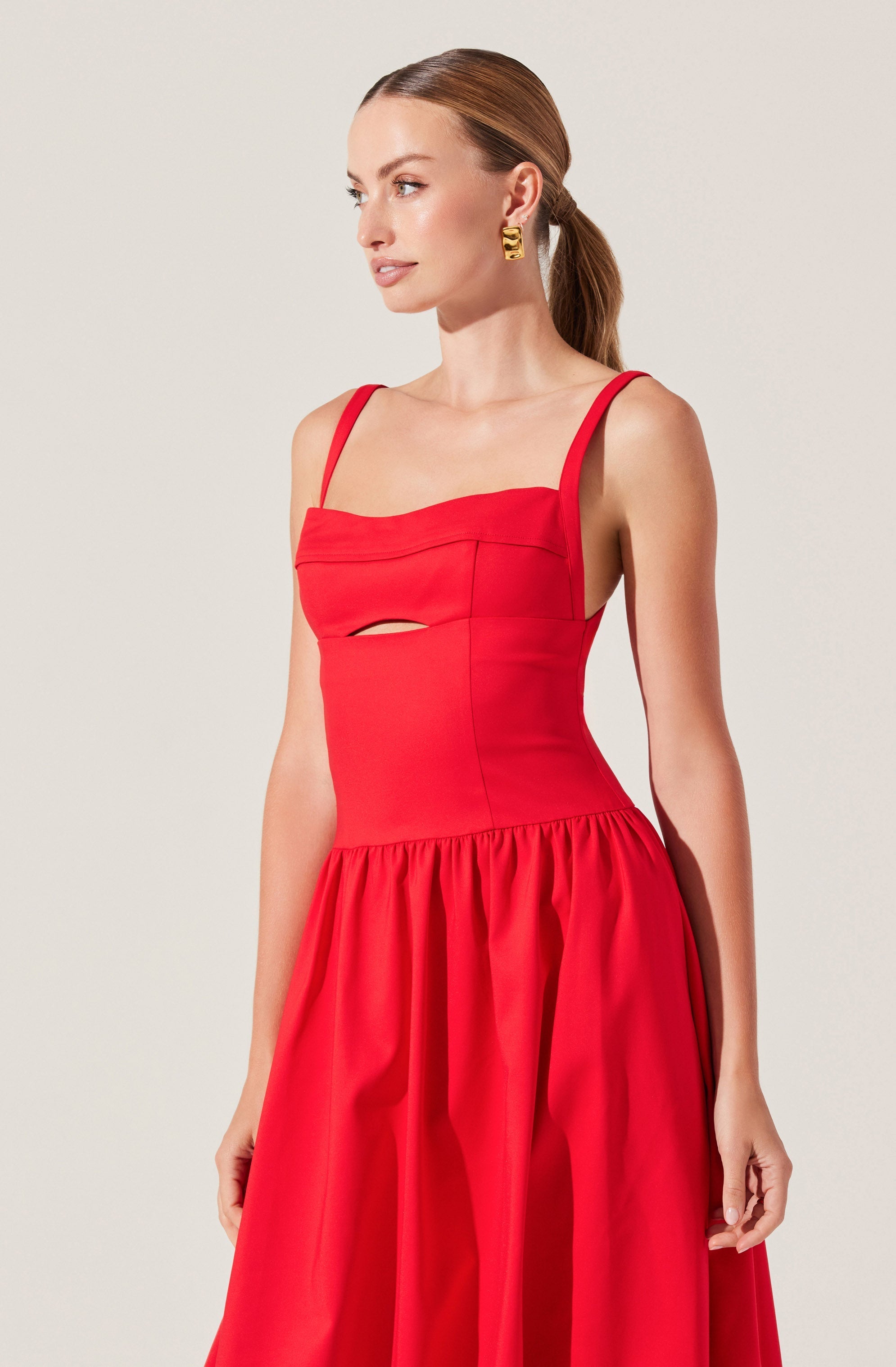 A woman is wearing a sleeveless, fitted red dress with thin shoulder straps. The dress features a small horizontal cutout detail just below the bust. The waistline is fitted, and the skirt is gathered, creating a full and voluminous silhouette. She has her hair pulled back and is accessorized with large, gold geometric earrings.