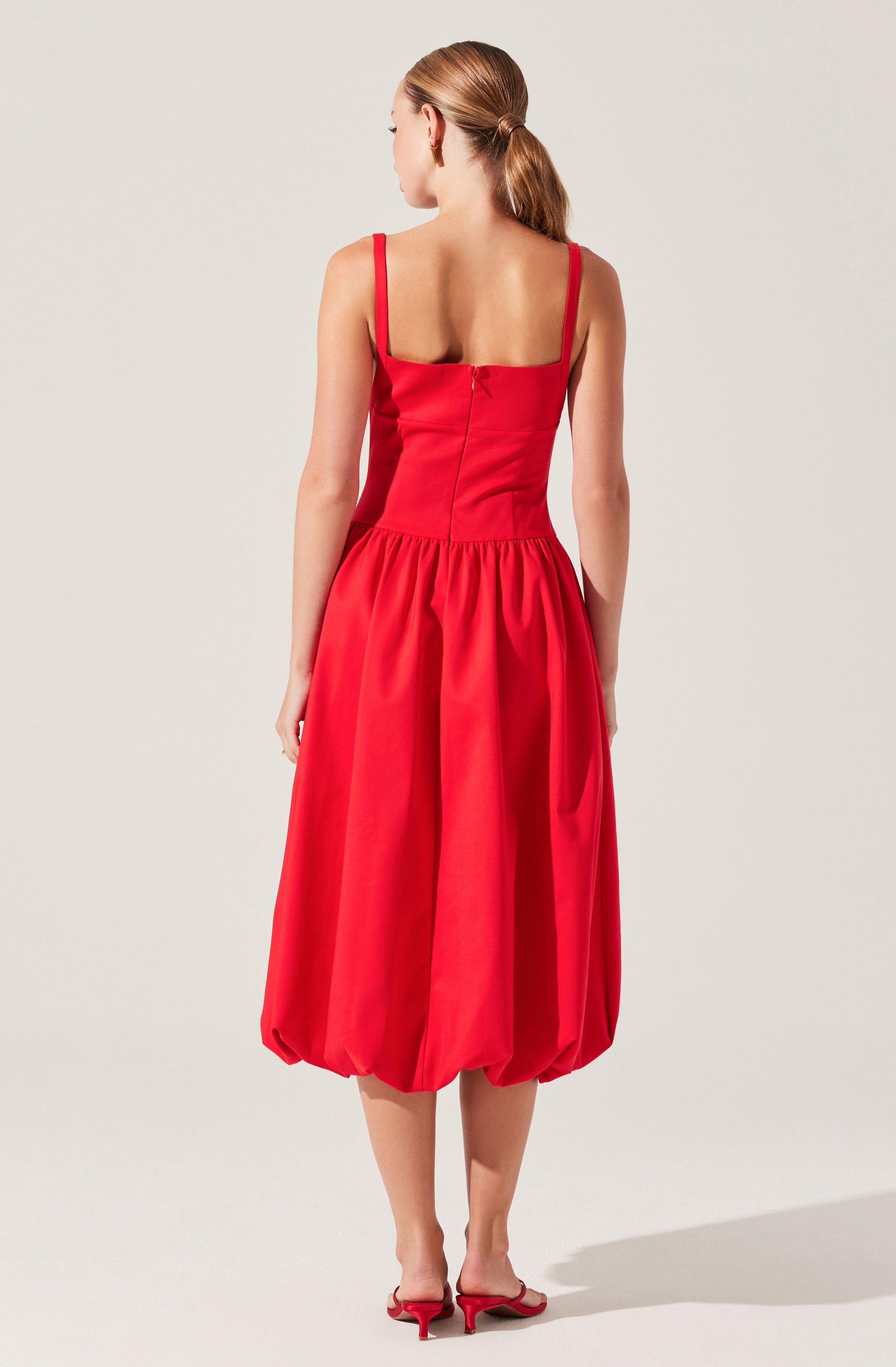 A woman is wearing a red dress with a fitted bodice and wide shoulder straps. The skirt is voluminous with gathered fabric and a balloon-style hem that falls below the knees. She is also wearing matching red heeled sandals.