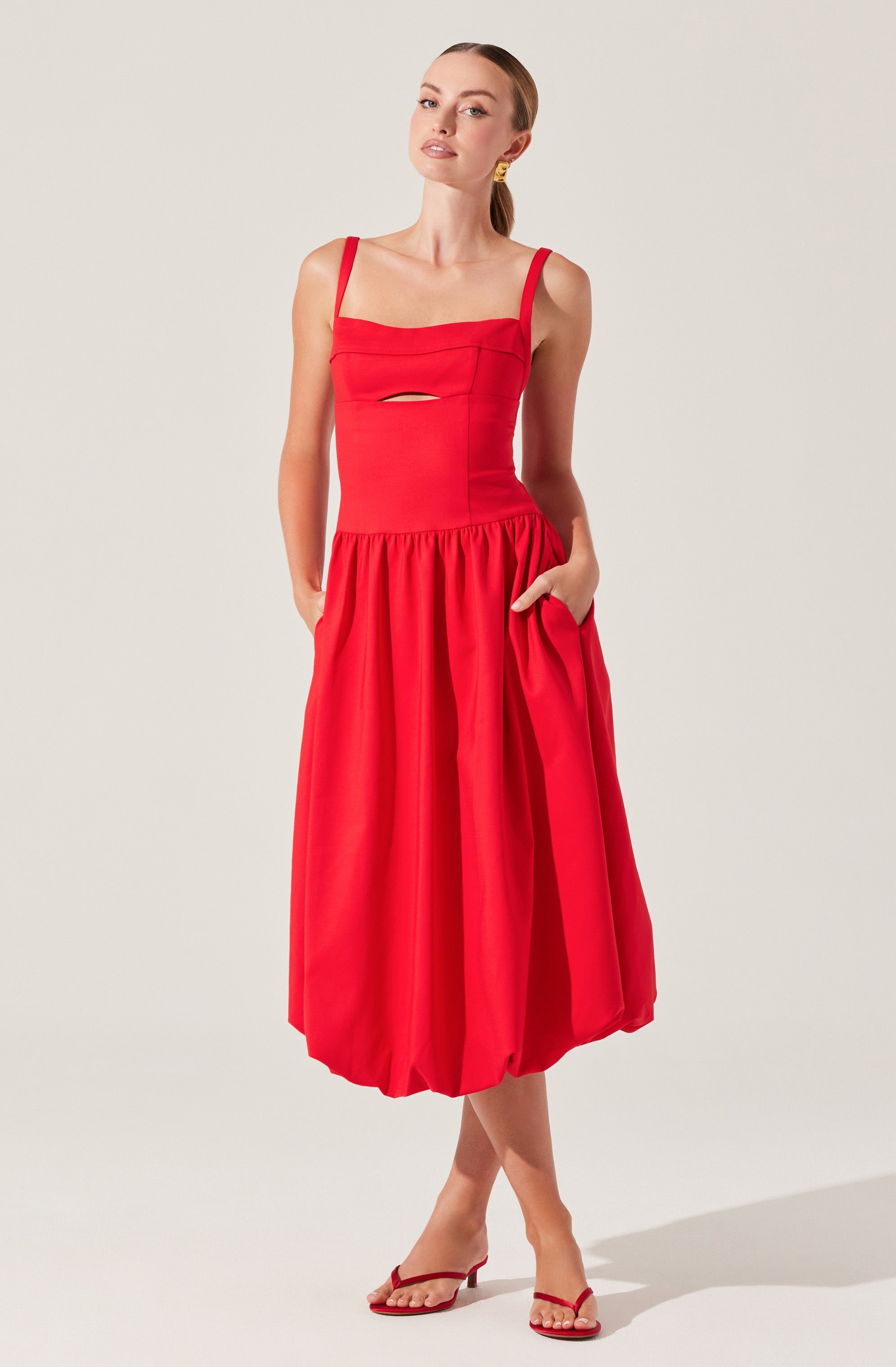 A woman is wearing a vibrant red sleeveless dress with adjustable shoulder straps. The fitted bodice features a small horizontal cutout just below the bust. The skirt is voluminous and gathered at the waist, reaching mid-calf length, and has a built-in pocket on the right side. She is also wearing matching red strappy sandals.