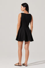 A woman is wearing a sleeveless black dress with a fitted bodice and a flared, pleated skirt that ends above the knees. She is also wearing black flat sandals and has gold earrings. Her dark hair is straight and shoulder-length.