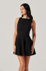 A woman is wearing a sleeveless black dress with a fitted bodice and a flared, pleated skirt. The dress has a round neckline and a subtle sheen, giving it an elegant appearance.