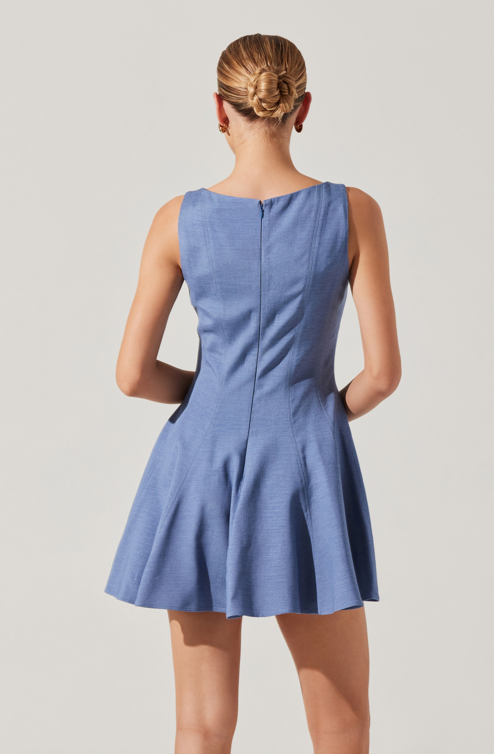 A woman is wearing a sleeveless, knee-length dress made of shiny blue fabric. The dress has a fitted bodice with a visible back zipper and a flared skirt with pleats.
