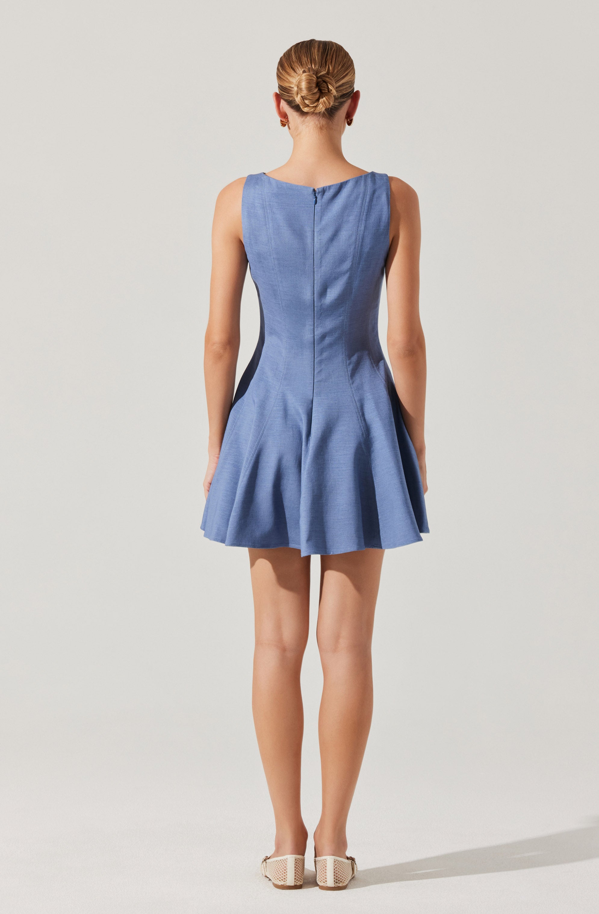 A woman is wearing a sleeveless, fitted blue dress with a flared skirt. The dress has a round neckline and appears to have dark side panels and a vertical back zipper. She is also wearing white shoes with a textured, possibly woven, pattern.