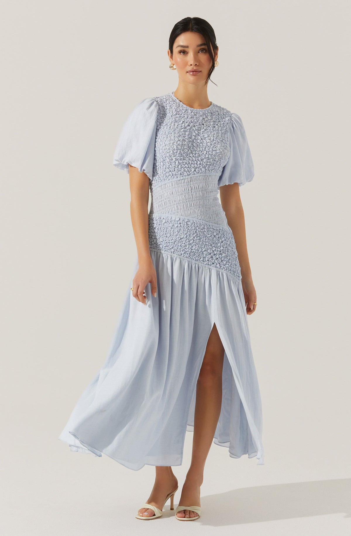 A woman is wearing a long, light blue dress with a fitted bodice and a flowing skirt. The dress features puffed short sleeves made of sheer fabric, with gathered details. The bodice has textured, ruched fabric with smocking, and the skirt has a high slit revealing one leg. The woman is also wearing beige high-heeled shoes.