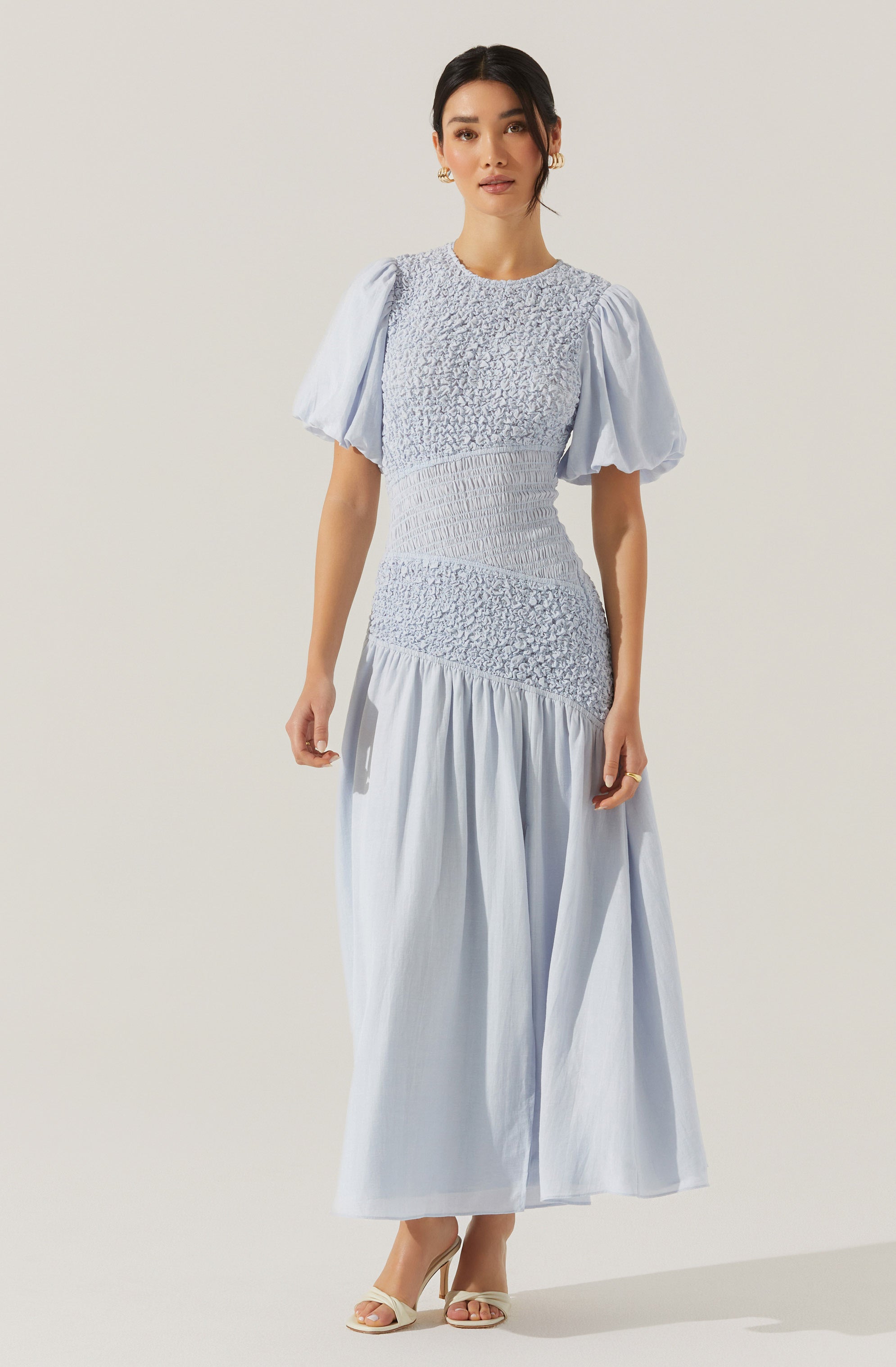 A woman is wearing a light blue dress with a fitted bodice featuring textured, gathered fabric. The dress has puffy short sleeves and a full-length skirt with a gathered, flowing design. She is also wearing open-toe heels.