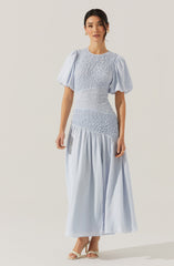 A woman is wearing a light blue dress with a fitted bodice featuring textured, gathered fabric. The dress has puffy short sleeves and a full-length skirt with a gathered, flowing design. She is also wearing open-toe heels.