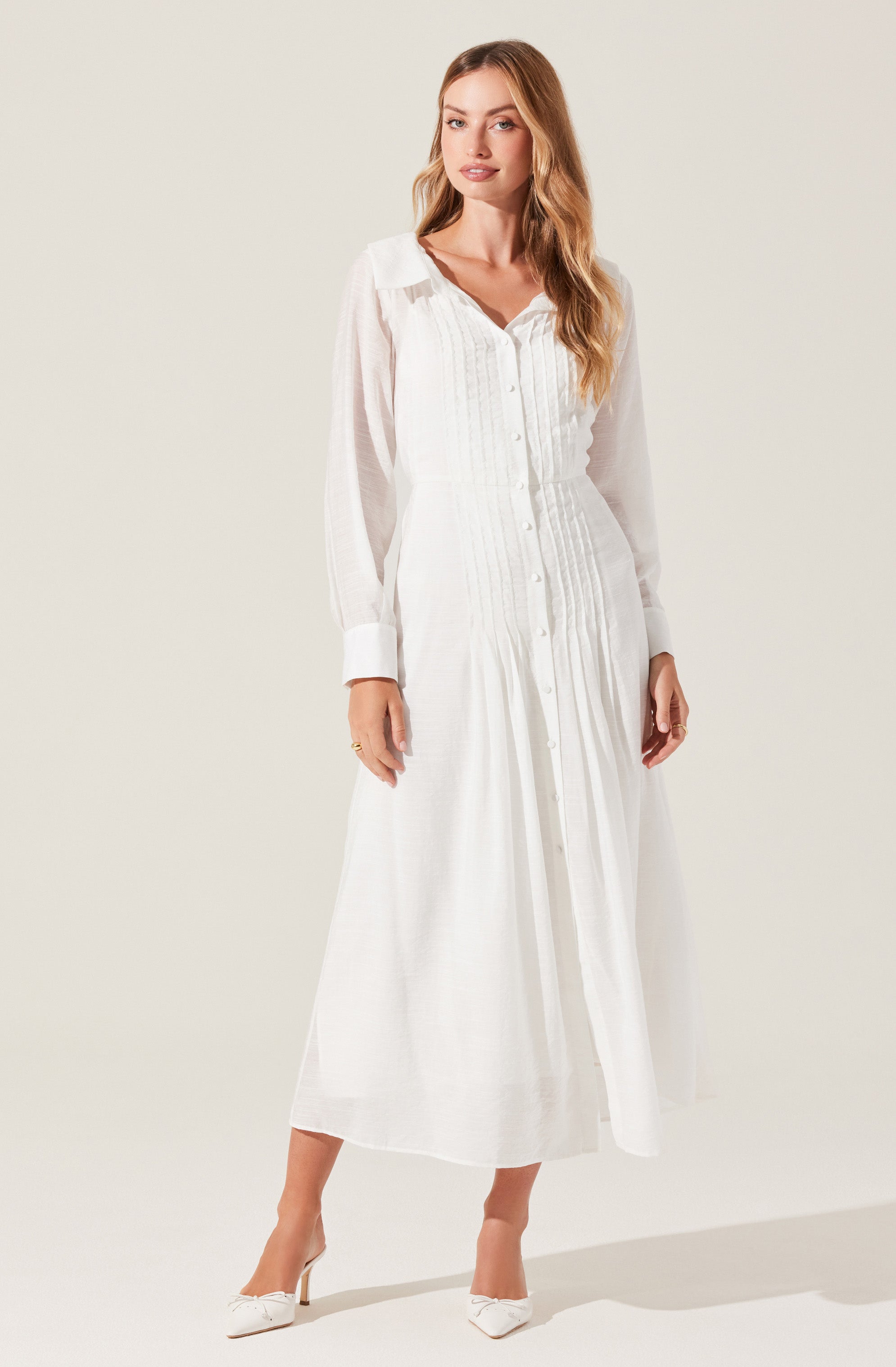 A woman is wearing a long, white, button-up dress with a collared neckline. The dress features vertical pleats and sheer, long sleeves with wide cuffs. She is also wearing white high-heeled shoes.
