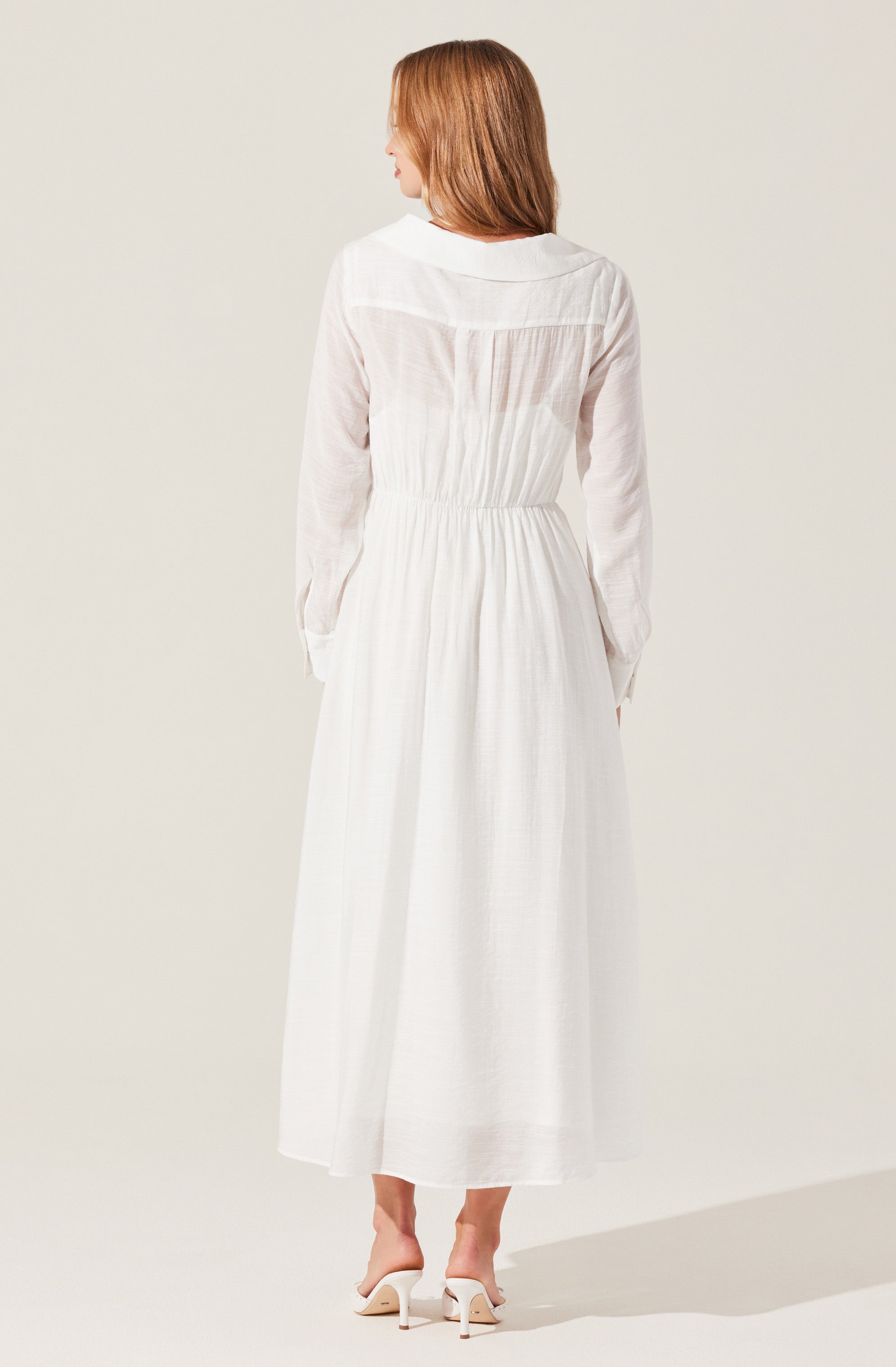 A woman is wearing a long, white, flowing dress with long sleeves. The dress has a fitted waist and a gathered skirt that reaches her ankles. The fabric appears lightweight and semi-sheer, giving it an airy, elegant look. She is also wearing a pair of white heeled sandals.