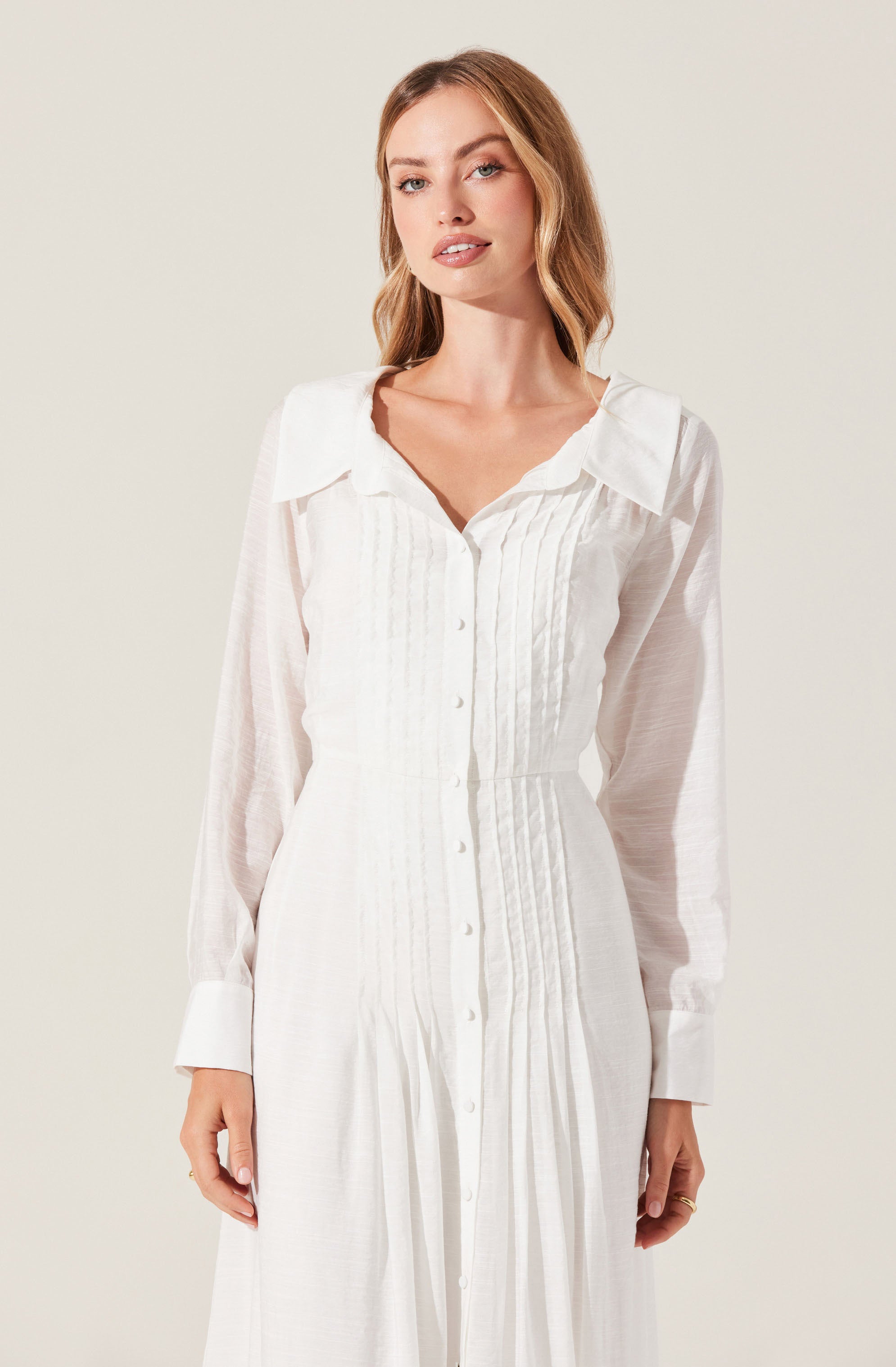 A woman is wearing a long, white, button-down dress with a collar. The dress features vertical pleats along the front and long sheer sleeves with wide cuffs.