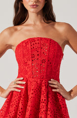 The woman is wearing a strapless red dress with intricate eyelet embroidery and cut-out floral patterns throughout the fabric. The dress features a fitted bodice and a flared skirt, creating a feminine and elegant silhouette.