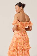 A woman is wearing an off-the-shoulder dress with ruffled sleeves. The dress features a blend of soft pastel colors, including shades of pink, orange, and yellow, with a pattern that appears to be a mix of tie-dye and vertical stripes. The fabric looks lightweight and flowy, and the dress has a tiered design with ruffles and pleats. She is accessorized with gold hoop earrings and has her dark hair styled in a sleek, low bun.