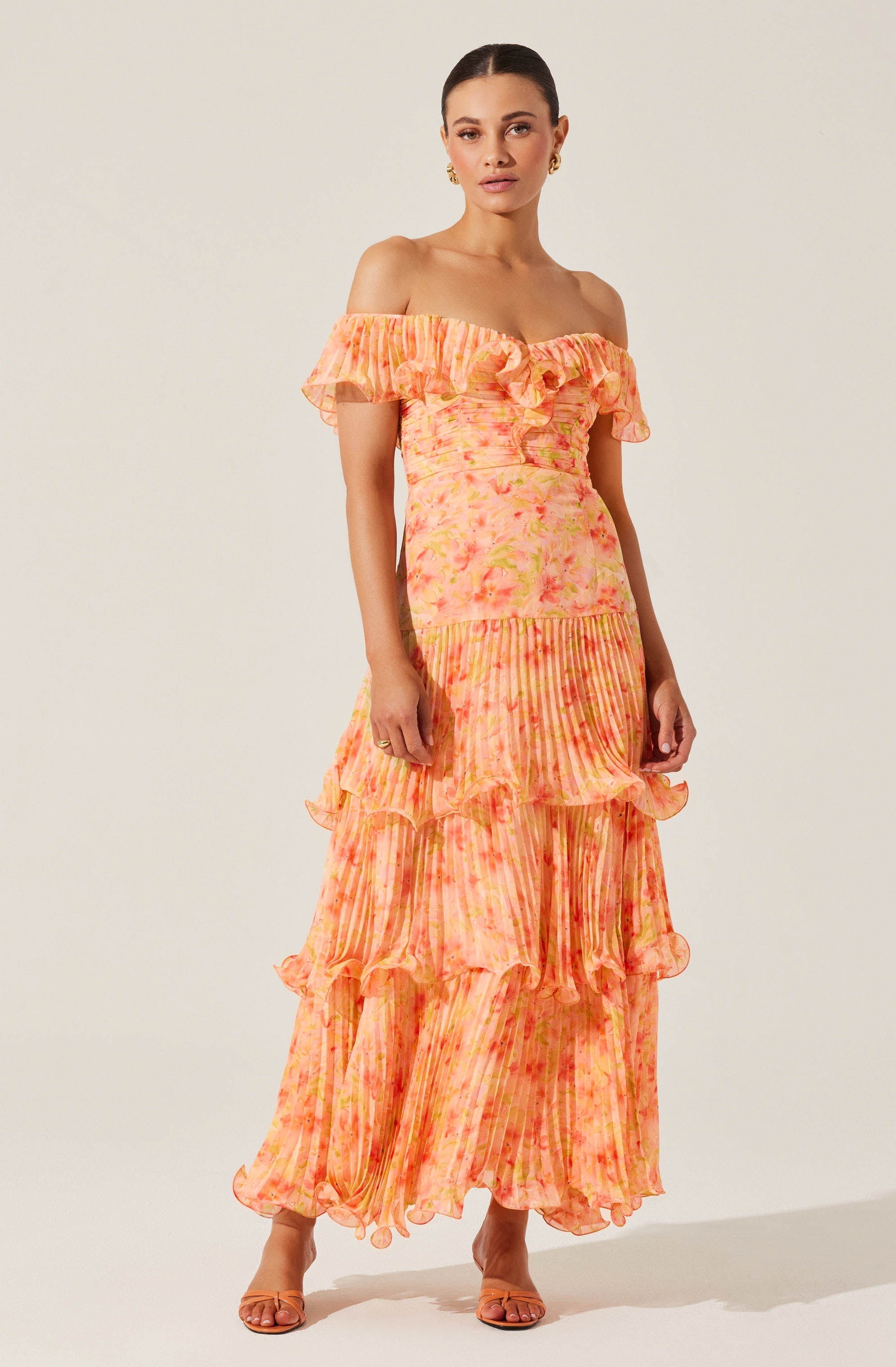 A woman is wearing a long, off-the-shoulder dress with a ruffled neckline. The dress features a colorful, tie-dye pattern in shades of pink, orange, and yellow, with a fitted bodice and tiered, ruffled layers extending to the floor. The fabric appears lightweight and flowy.