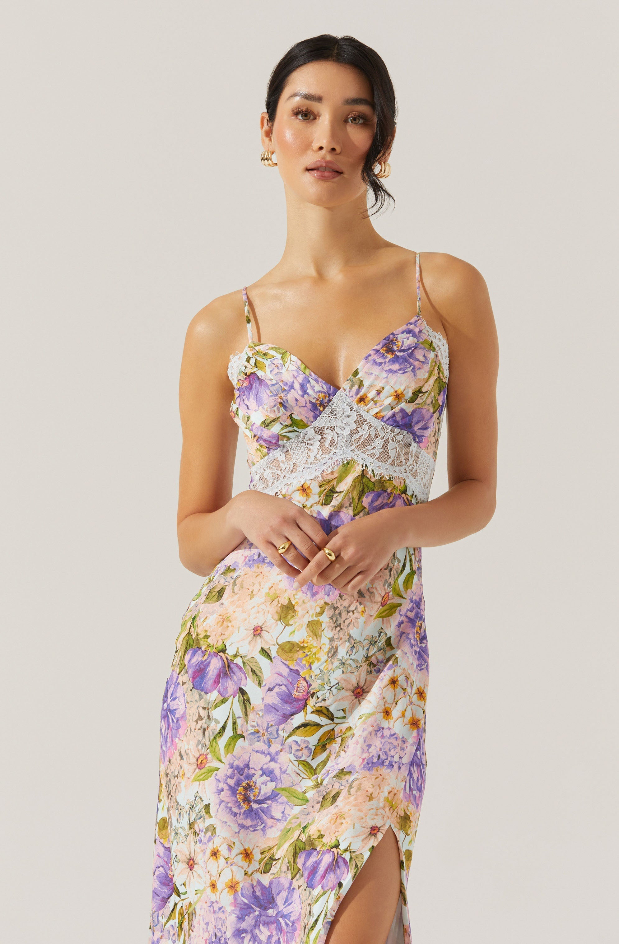 A woman is wearing a sleeveless, floral dress with spaghetti straps. The dress features a vibrant pattern of purple, lavender, pink, and yellow flowers with green leaves on a light background. The top of the dress has a lace detail under the bust, adding a delicate touch. The skirt has a slit on one side, showing a bit of leg.