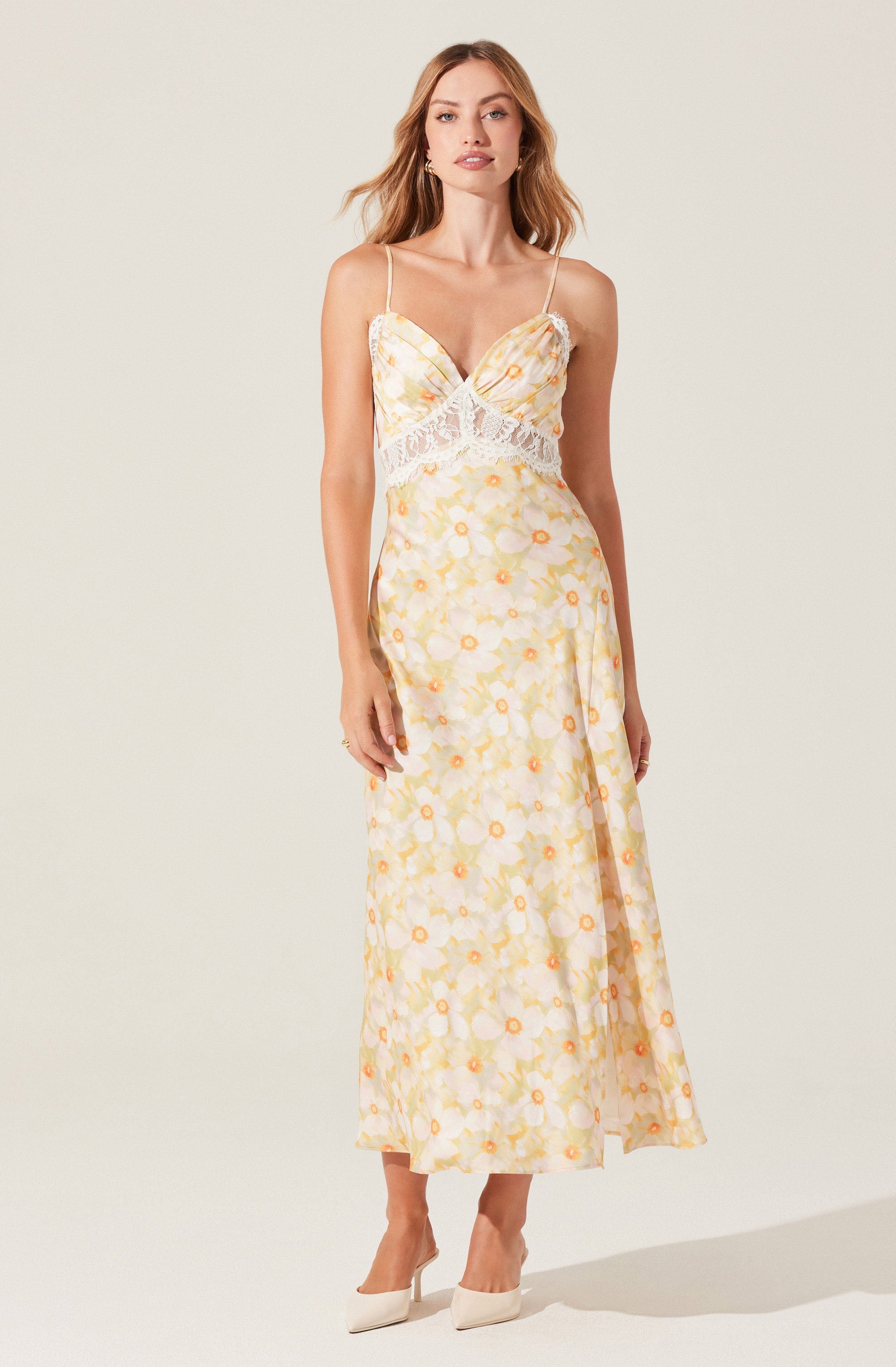 A woman is wearing a light-colored, sleeveless maxi dress with thin spaghetti straps. The dress features a floral pattern in shades of yellow and orange with some green accents. The bodice has a ruched detail, and there is a white lace trim around the waist, adding a decorative touch. She is also wearing white high-heeled shoes.