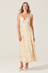 A woman is wearing a light-colored, sleeveless maxi dress with thin spaghetti straps. The dress features a floral pattern in shades of yellow and orange with some green accents. The bodice has a ruched detail, and there is a white lace trim around the waist, adding a decorative touch. She is also wearing white high-heeled shoes.