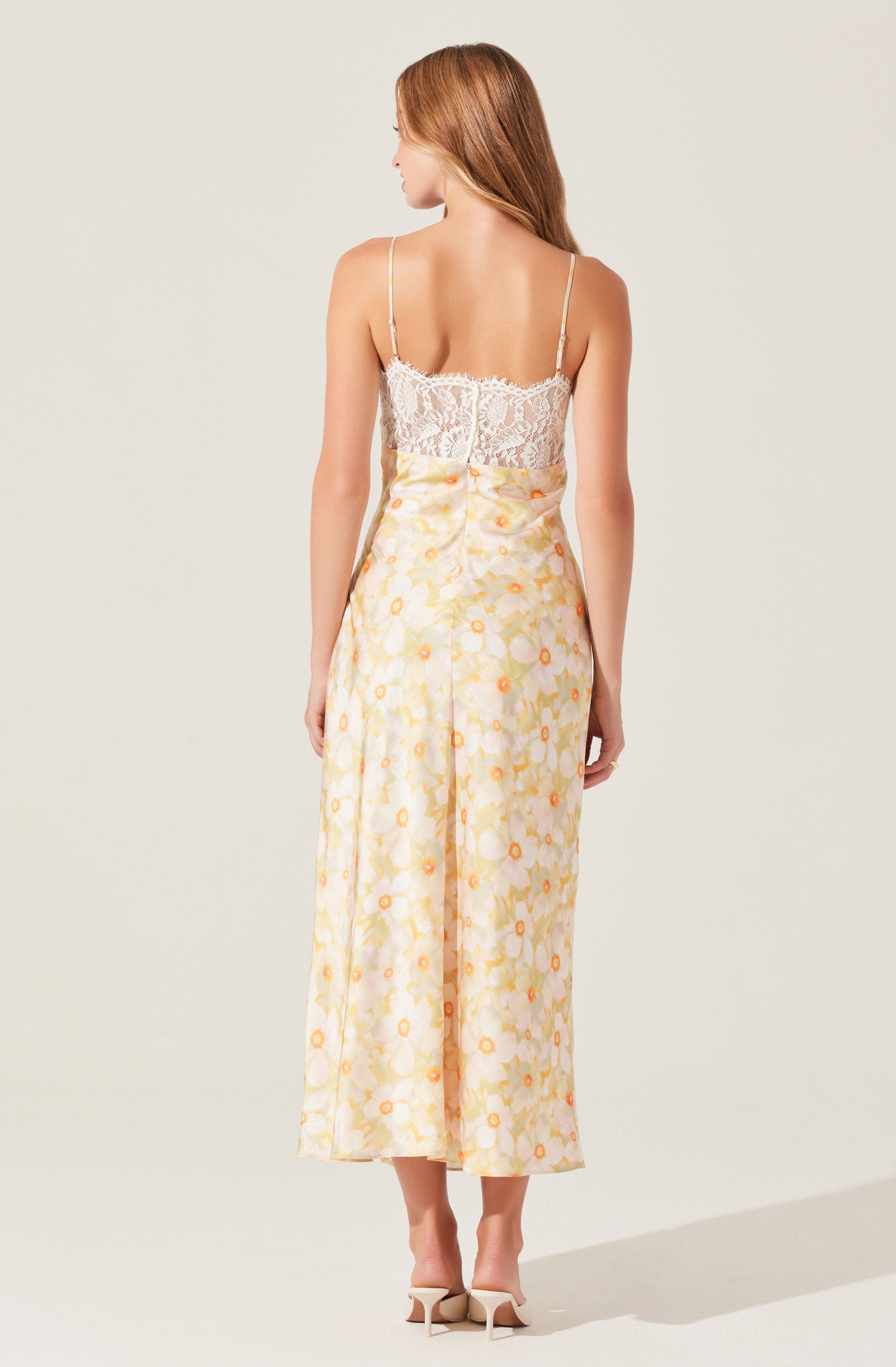 A woman is wearing a sleeveless, fitted top made of white lace fabric with delicate spaghetti straps. She is also wearing high-waisted, wide-leg pants with a light yellow base color and a floral pattern featuring orange and yellow flowers.