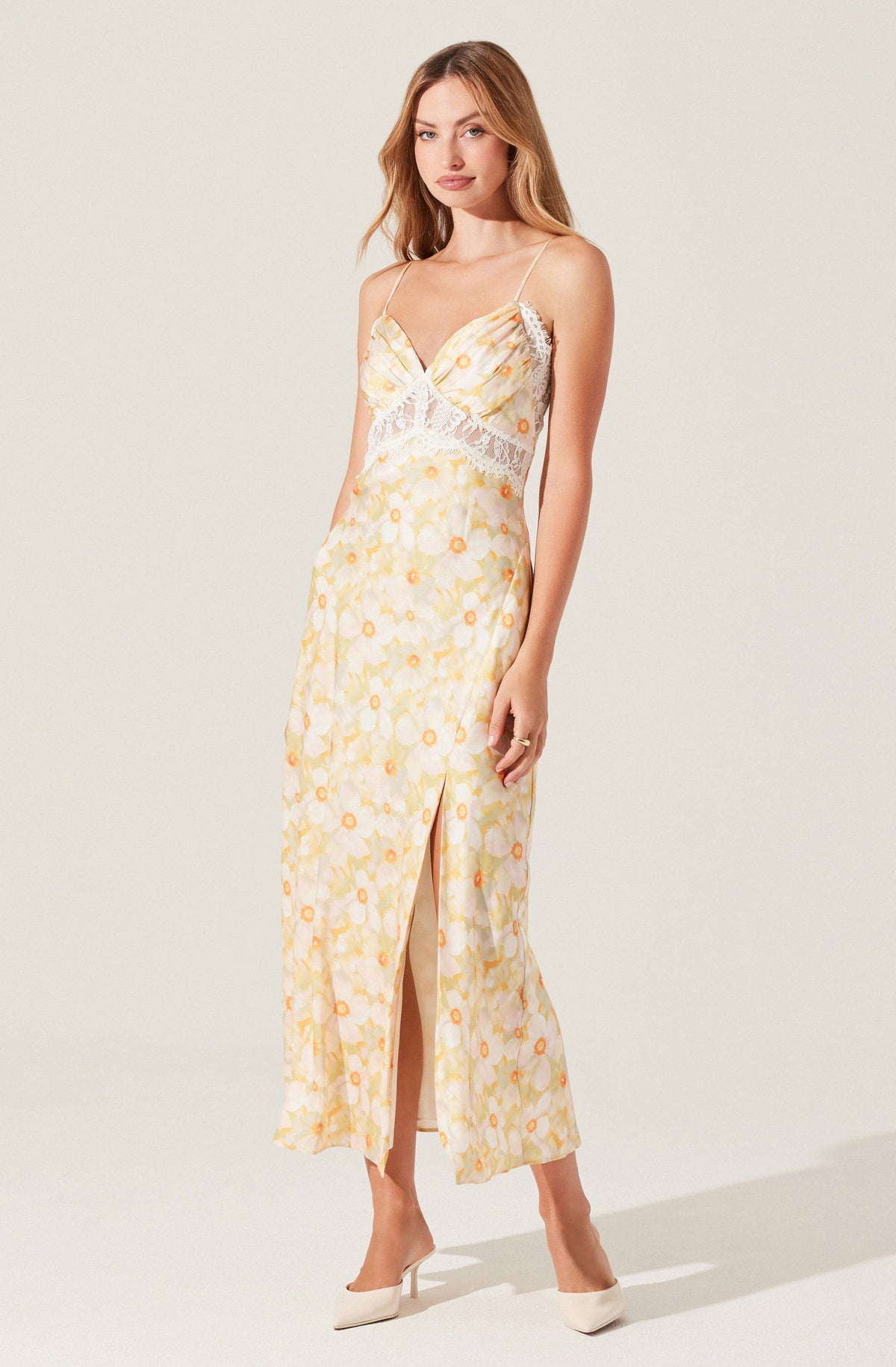 A woman is wearing a sleeveless, full-length dress with thin spaghetti straps. The dress has a light, semi-sheer fabric with a yellow and white floral pattern, and features lace detailing around the waist. It has a front slit that extends above the ankle. She is also wearing white high-heeled shoes.