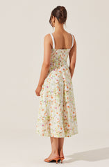 A woman is wearing a sleeveless, knee-length dress with a fitted bodice and a full skirt. The dress features wide straps with white lace trim and is made of a light, floral-printed fabric with pastel colors, including pink, orange, green, and white. The overall style is feminine and summery. The woman is also wearing open-toed high heels and has her hair styled in an elegant updo.