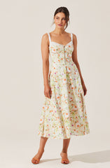 A woman is wearing a sleeveless, ankle-length dress with a floral pattern in pastel colors. The dress has wide, textured straps and a gathered neckline with a small bow at the center. The fabric features a light background with a variety of soft, colorful flowers. She is also wearing tan, open-toe heels.