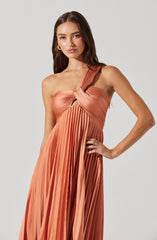 A woman is wearing a strapless, pleated, floor-length dress in a warm, peachy-orange color. The fabric is gathered and knotted at the chest, with one pleated strap over the shoulder. The pleats extend down the entire length of the dress, creating a flowing silhouette.