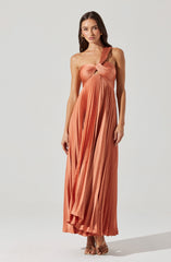 A woman is wearing a floor-length, pleated, rust-colored dress with a one-shoulder design. The dress features a twisted knot detail at the bust and has an asymmetrical strap on one shoulder, while the other shoulder is bare. The fabric appears shiny, adding a smooth texture to the pleats. She is also wearing open-toe high-heeled sandals.