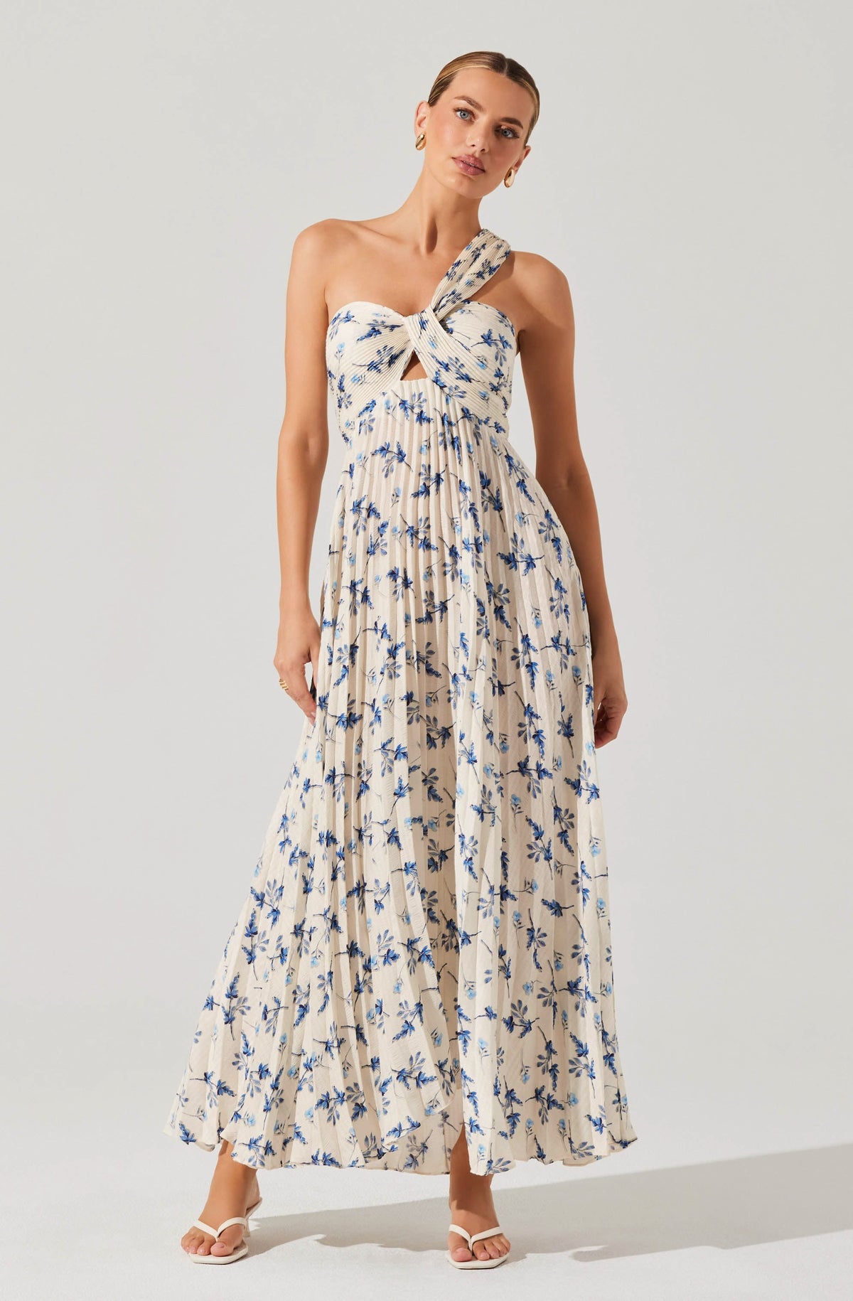A woman is wearing a sleeveless, long maxi dress with a light beige background and a blue floral print. The dress has a distinctive twisted knot detail at the bust with a small keyhole opening just below it. The skirt is flowing and pleated, creating a soft, airy silhouette.