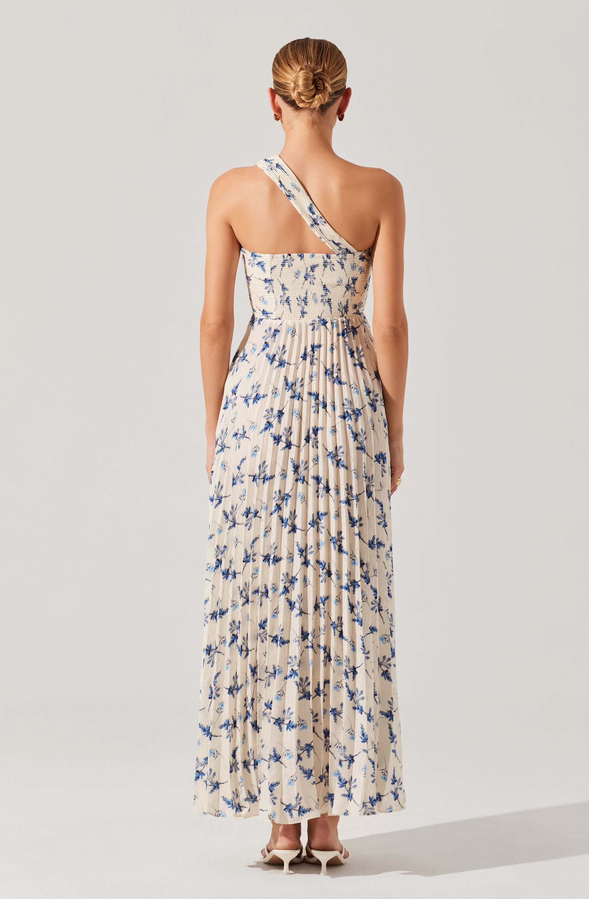 A woman is wearing a long, flowing dress with a beige base color and a blue floral print. The dress features a one-shoulder strap design. The fabric appears lightweight and comfortable, suitable for warm weather. She is also wearing neutral-colored high heels.