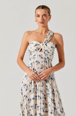 A woman is wearing a sleeveless, beige dress with a blue floral print. The dress features a large bow at the top, with one strap over her left shoulder and a small cutout below the bow. The fabric is pleated, creating a flowing appearance. She accessorized with gold hoop earrings and a ring on her right hand.
