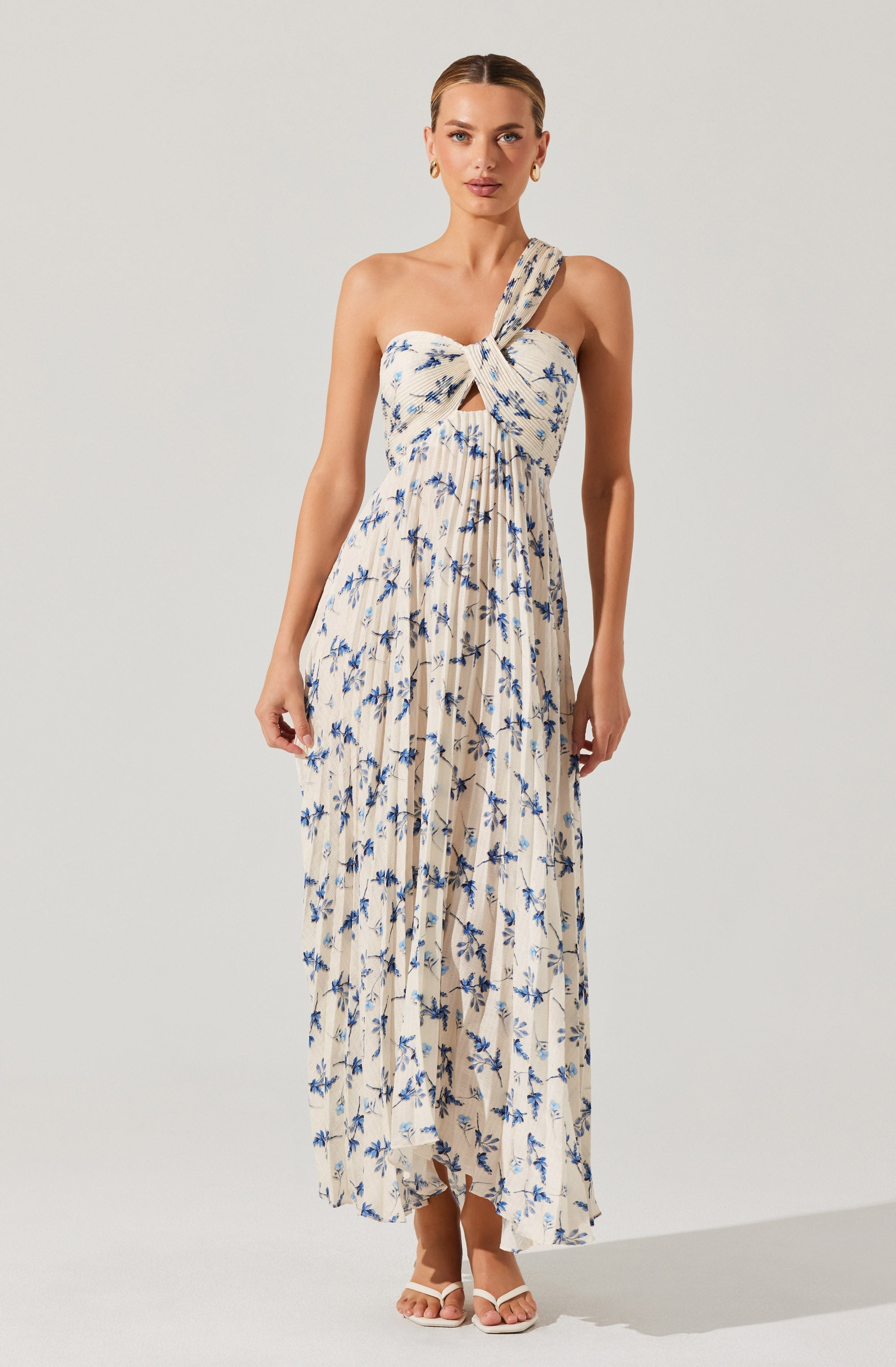 A woman is wearing a sleeveless, strapless maxi dress with a large bow at the front. The dress features a beige base color with a blue floral pattern, and it has a cut-out detail just below the bow. The fabric is lightweight and pleated, flowing down to her ankles. She is also wearing white heeled sandals.