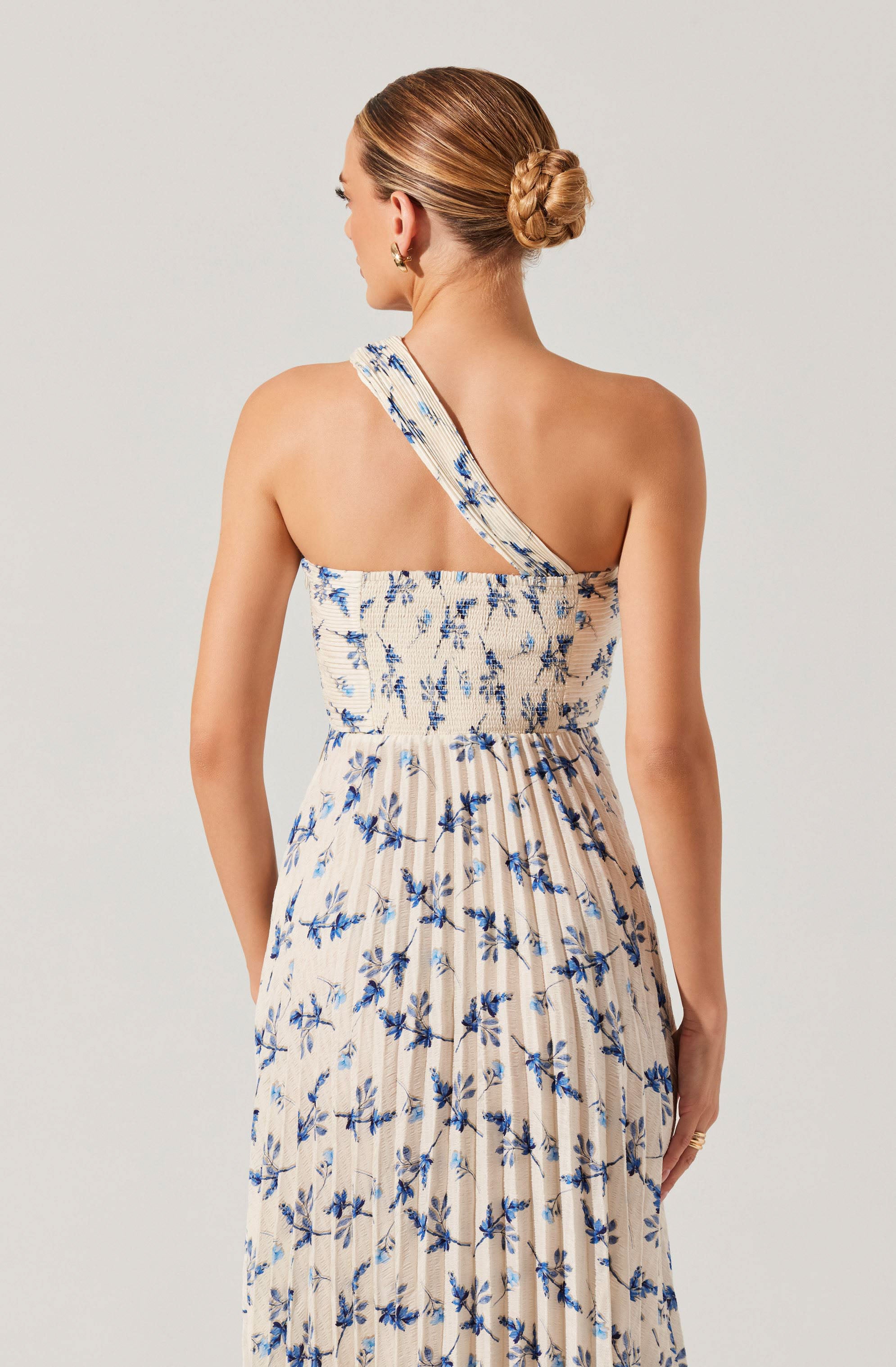 A woman is wearing a sleeveless, one-shoulder dress with a wide strap over her left shoulder. The dress features a light background color with a blue floral pattern and has a gathered, elastic waistband. The skirt portion of the dress is pleated and flows down to the floor. Her hair is styled in a neat, low bun.