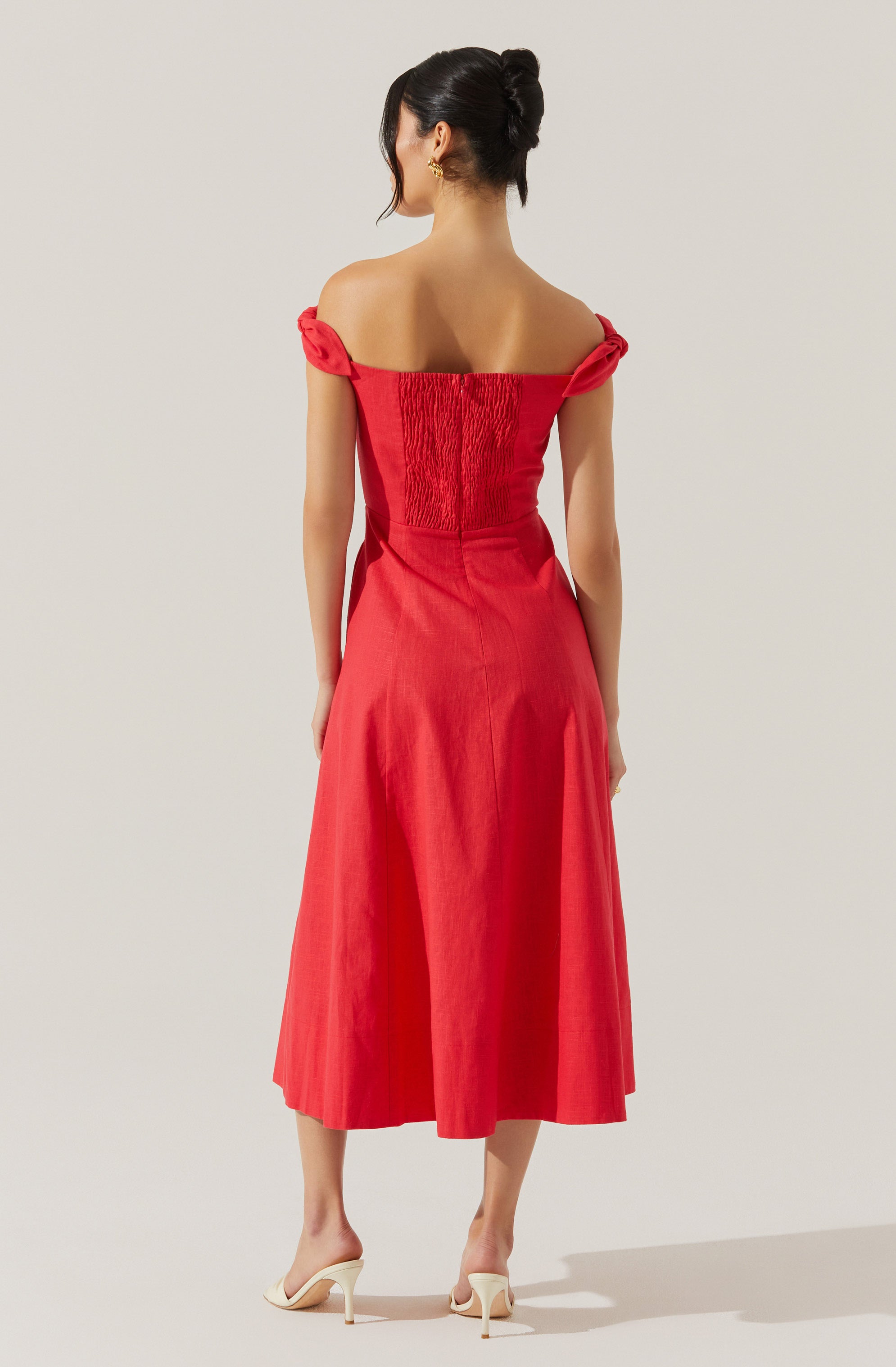 A woman is wearing a red off-the-shoulder dress with ruched detailing on the bodice and short, tied sleeves. The dress has a fitted waist and a flowy, mid-length skirt that reaches mid-calf. She is also wearing white high-heeled shoes.