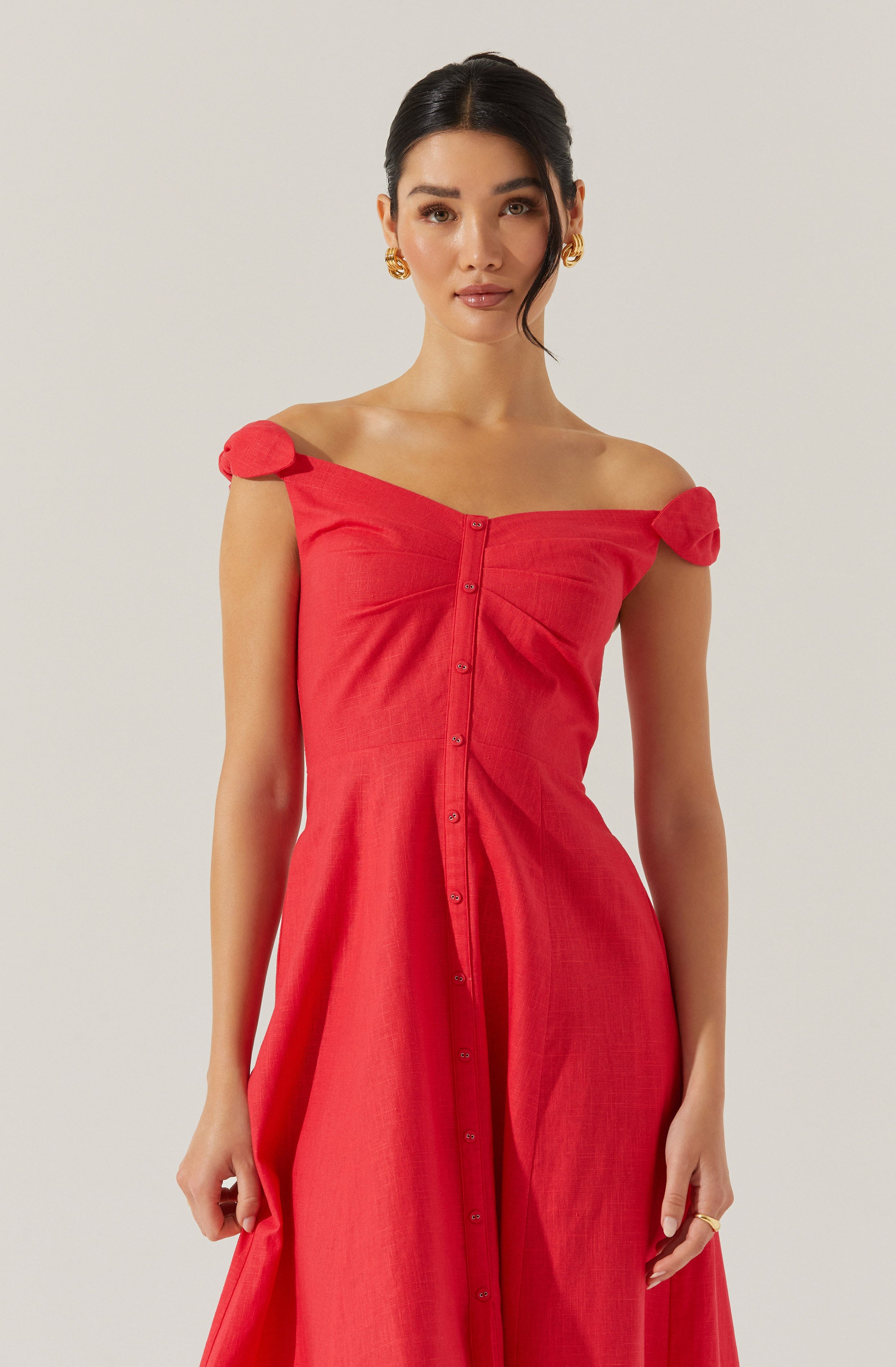 A woman is wearing a red, off-the-shoulder dress with short sleeves that have decorative fabric bows. The dress features a row of buttons running down the front. The fabric appears lightweight and slightly textured.