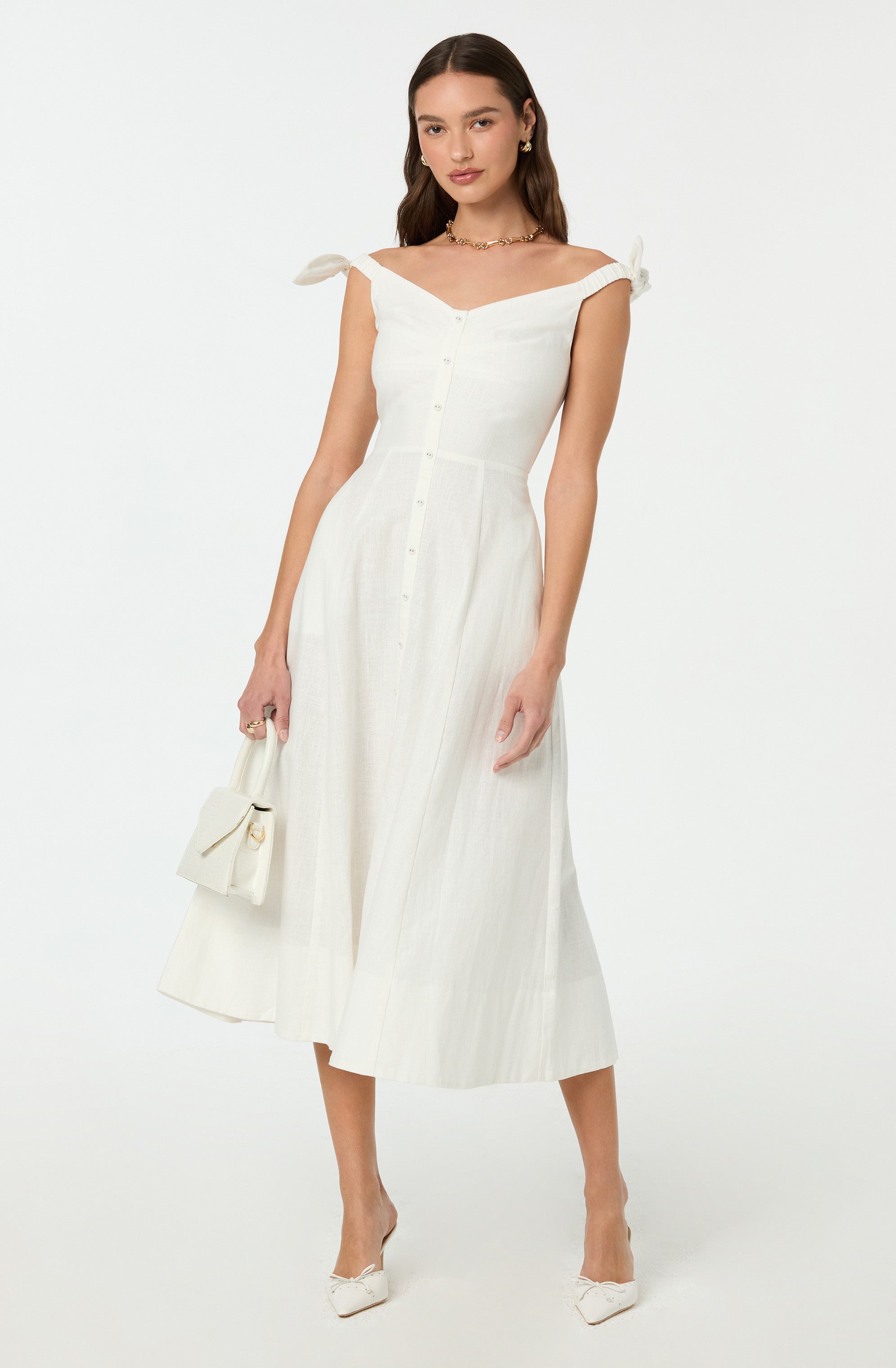 A woman is wearing a white, sleeveless dress with tied shoulder straps and a row of buttons down the front. She is holding a matching white handbag and wearing white high-heeled shoes.