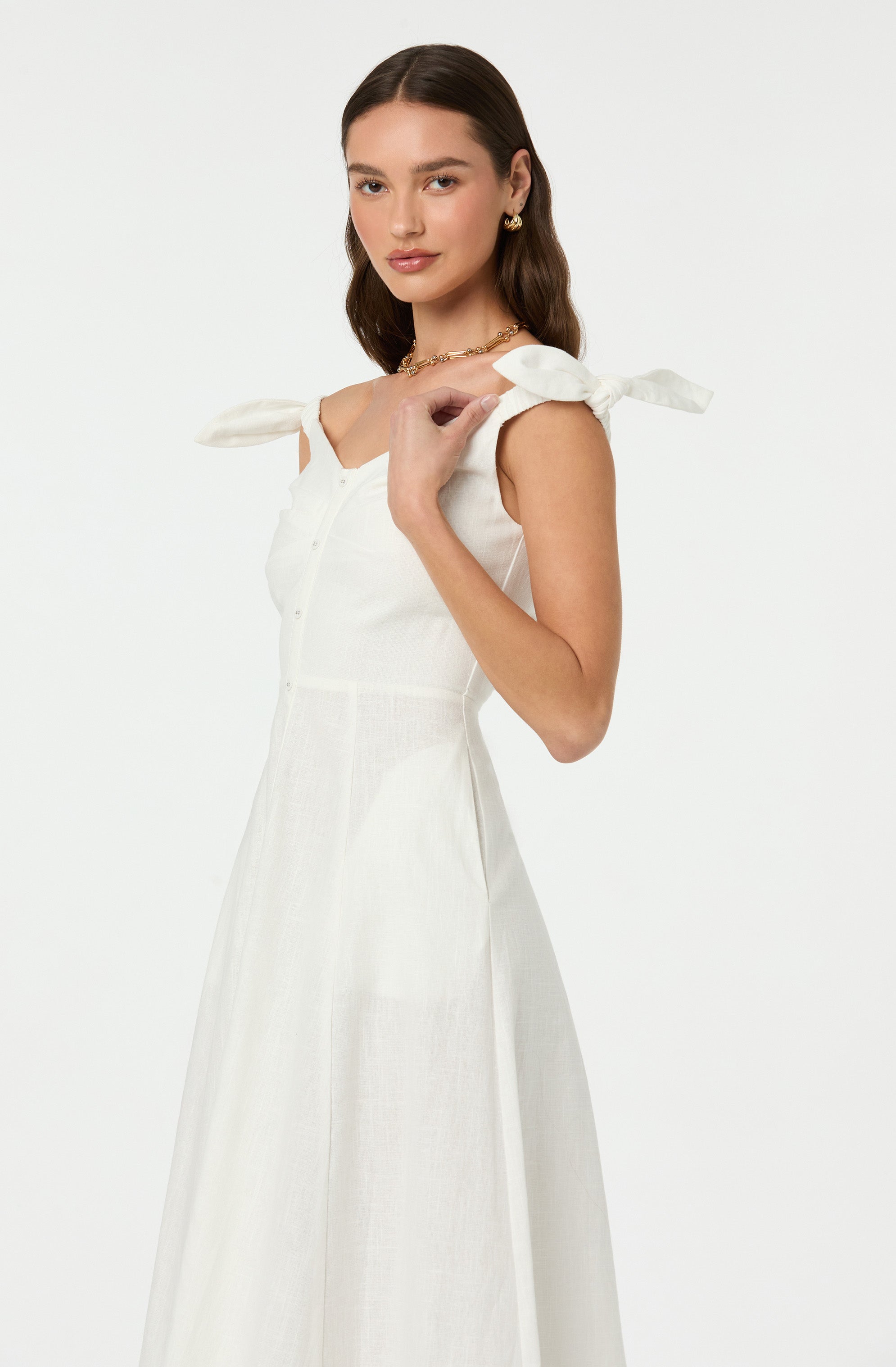 A woman is wearing a sleeveless white dress with bow-tied shoulder straps. The dress features a button-down front and a fitted waist, flowing into a long, elegant skirt. She accessorizes with gold earrings and a gold necklace.