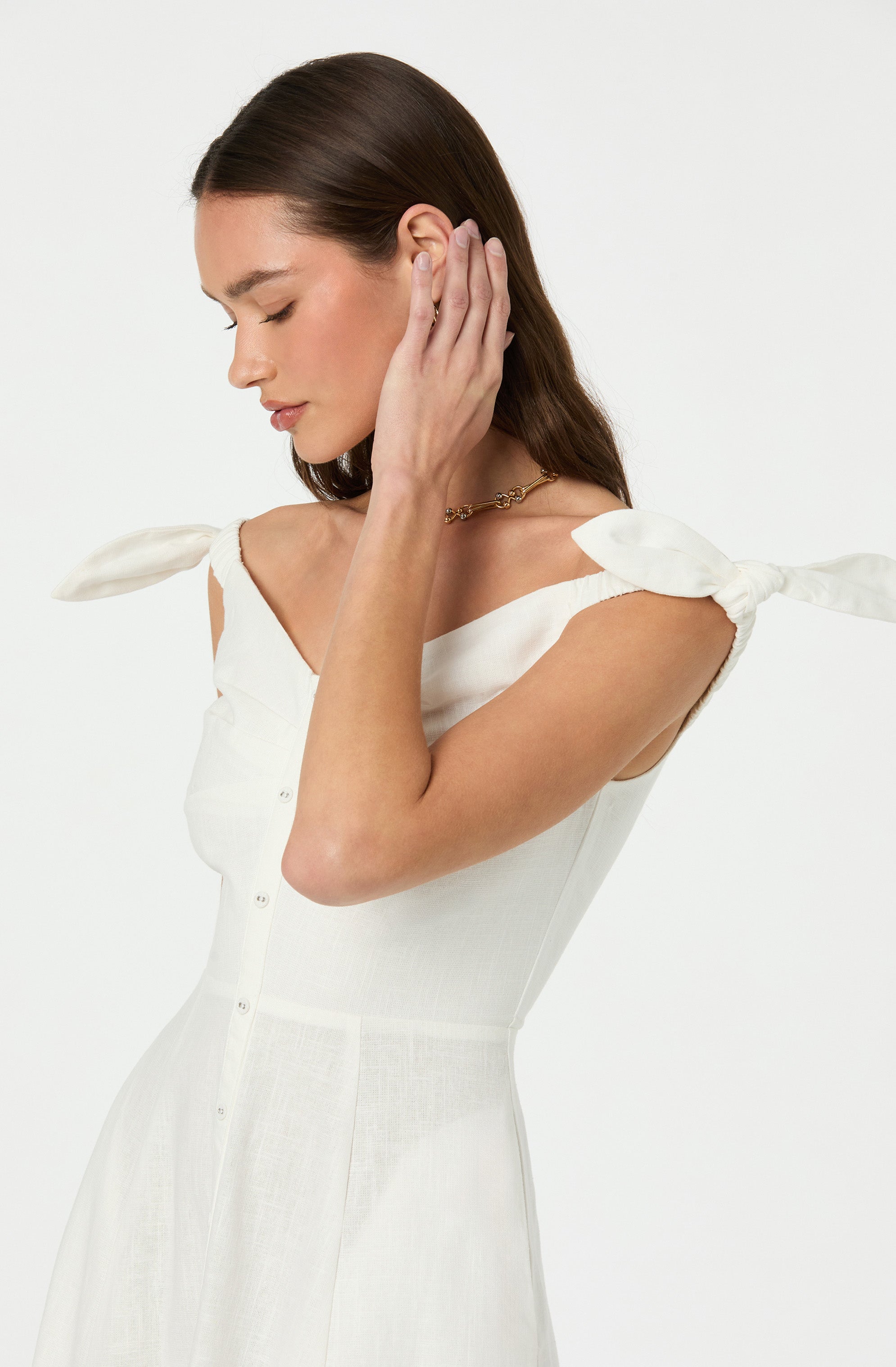 A woman wearing a white sleeveless dress with shoulder ties and a v-neckline. The dress has a fitted waist and buttons running down the front. She is also wearing a delicate gold necklace.