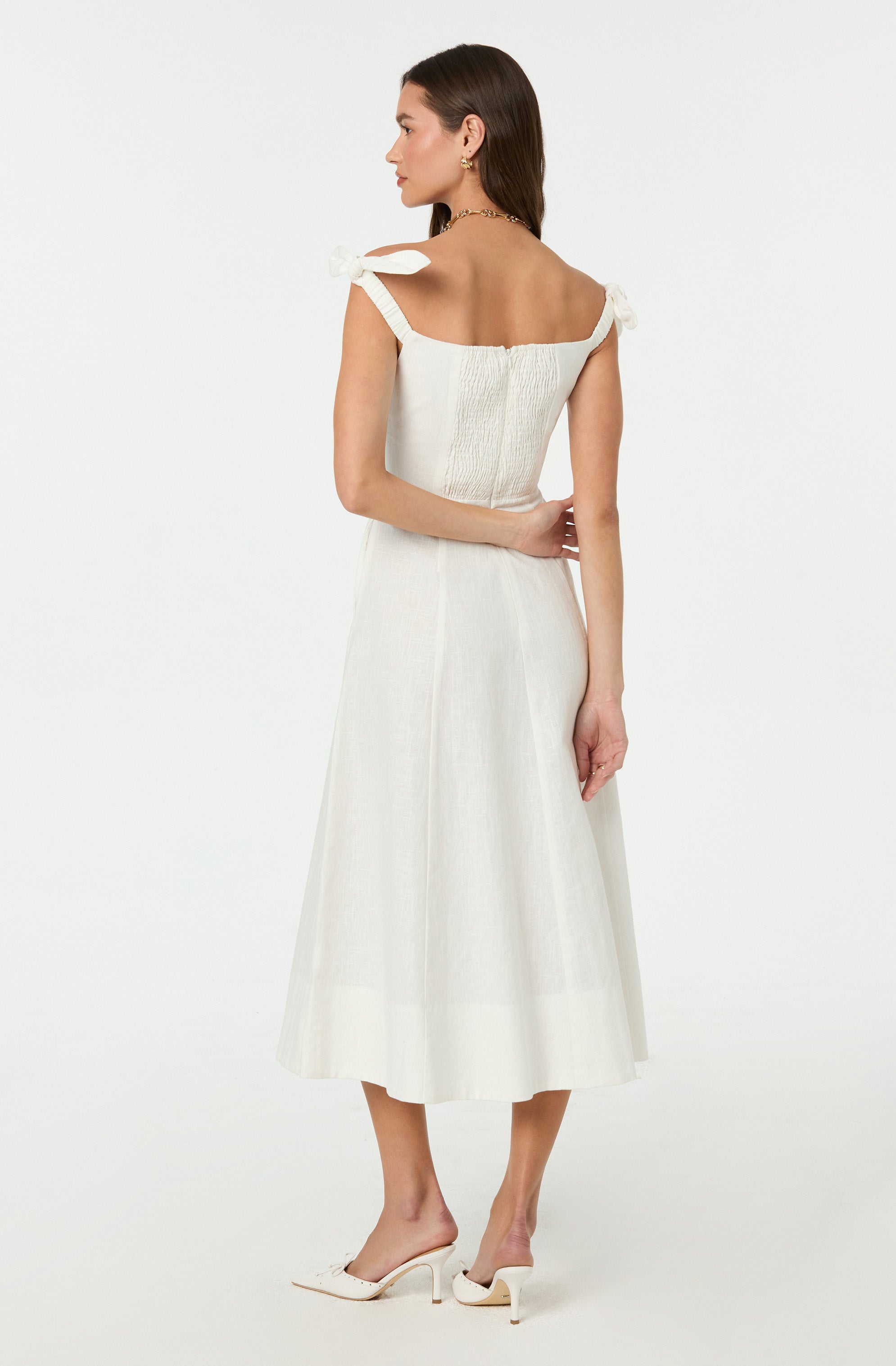 A woman is wearing a sleeveless, off-white dress with shoulder straps tied into bows. The top has a textured, ruched design, and the skirt flows down to mid-calf length. She pairs the dress with white, open-toed high heels.