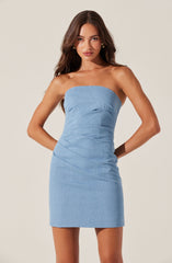 A young woman is wearing a strapless, light blue denim dress that is fitted and ends above the knees. She has long, wavy brown hair and is accessorized with gold earrings. The dress features a simple design with subtle ruching details.