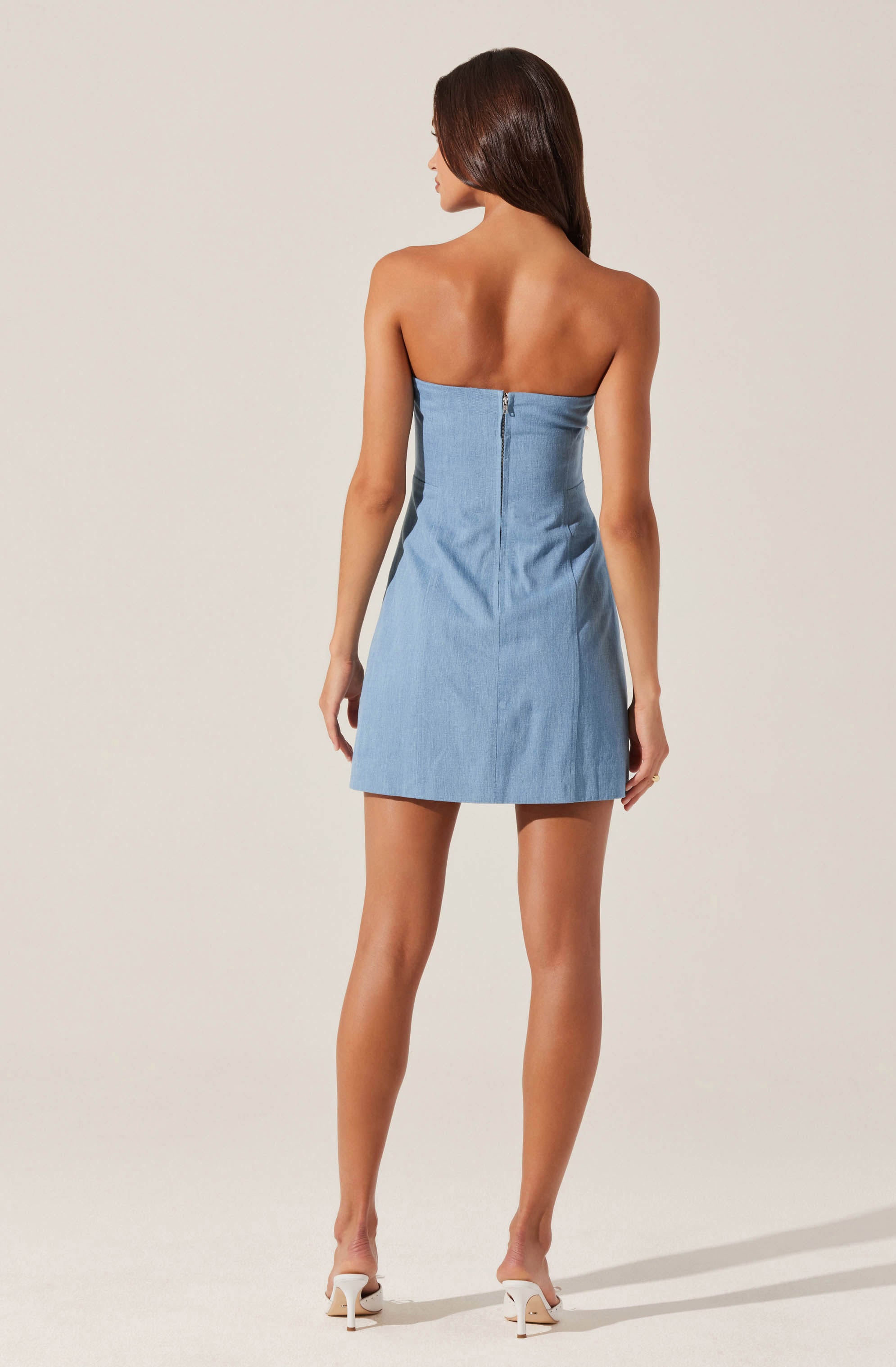 A woman is wearing a strapless, light blue denim mini dress with a visible back zipper closure. She has nude high heels and long, dark brown hair.