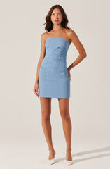 The dress is a strapless, light blue mini dress with a fitted silhouette that features subtle ruching details. The fabric appears to be a textured material, giving it a casual yet elegant look. The model pairs the dress with white pointed-toe heels.