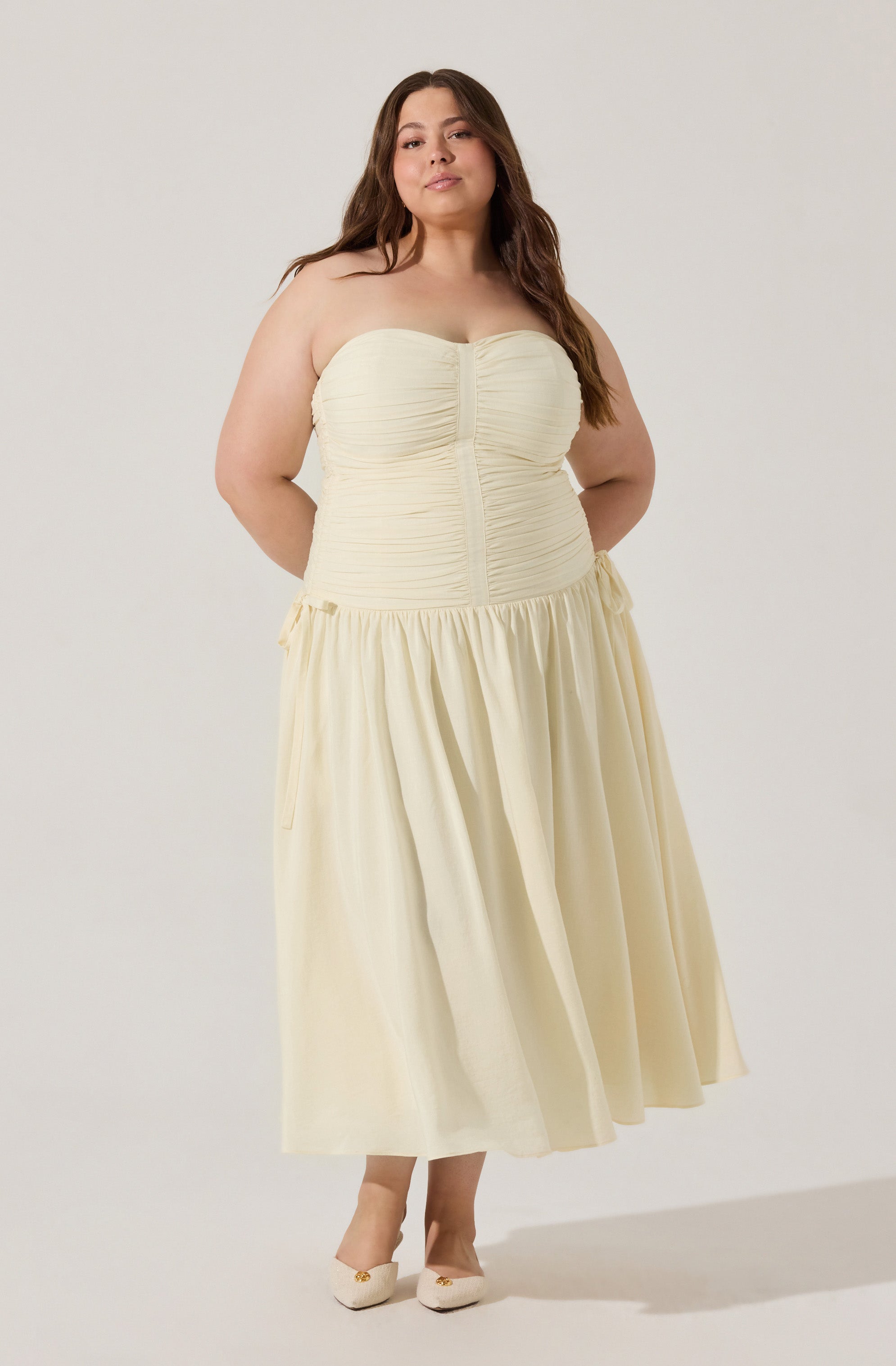 A woman is wearing a strapless, cream-colored dress with a ruched bodice and a gathered, flowing skirt that falls to her ankles. She has on matching cream-colored heels.